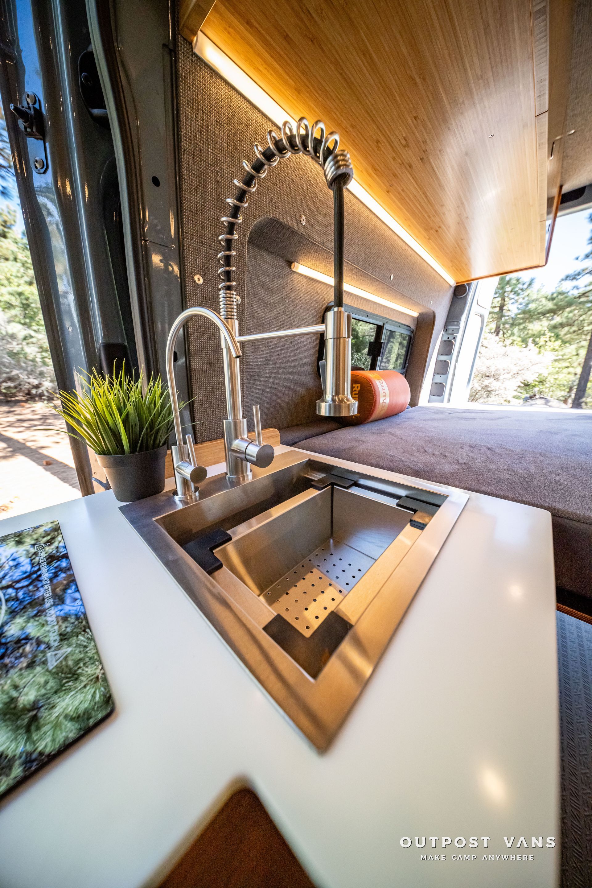 Sprinter Interior of a van with a stainless steel sink and faucet; a bed and counter space with a plant are visible.
