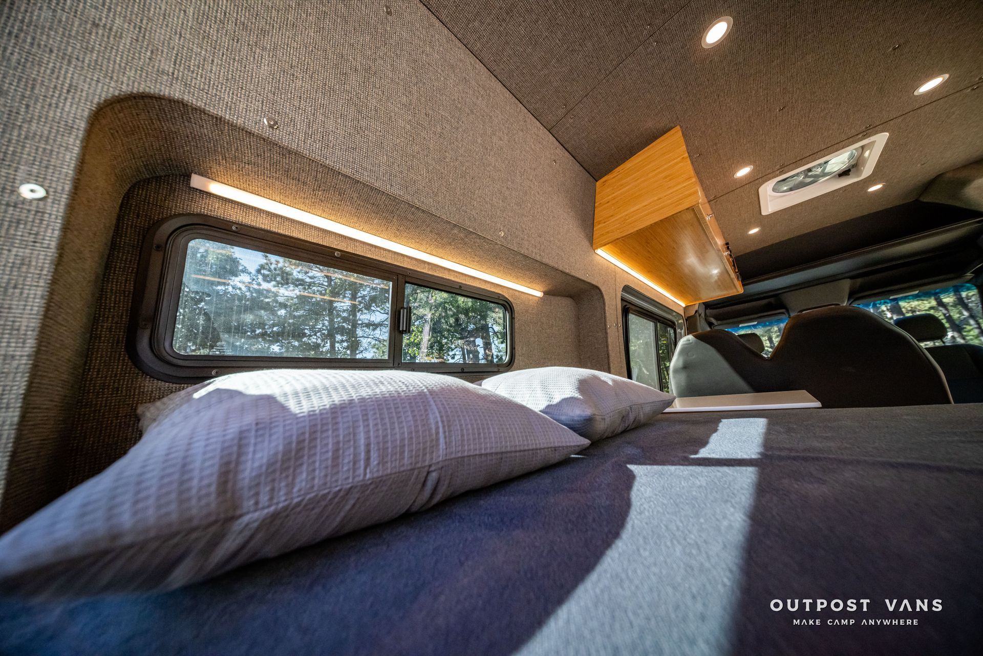 Sprinter Cozy van interior with bed, pillows, window, and overhead lighting. Grey and wood tones. Sunlight streams in.