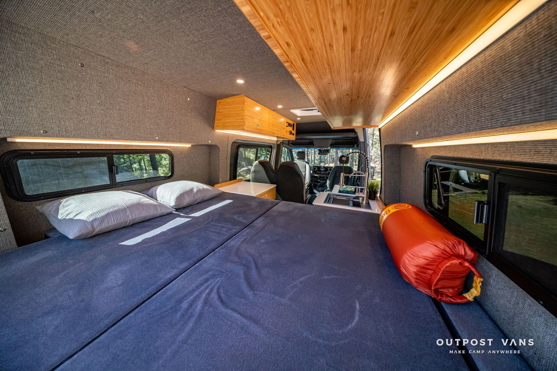 Sprinter Interior of a camper van with a bed, wood paneling, and windows. Red sleeping bag on the bed.