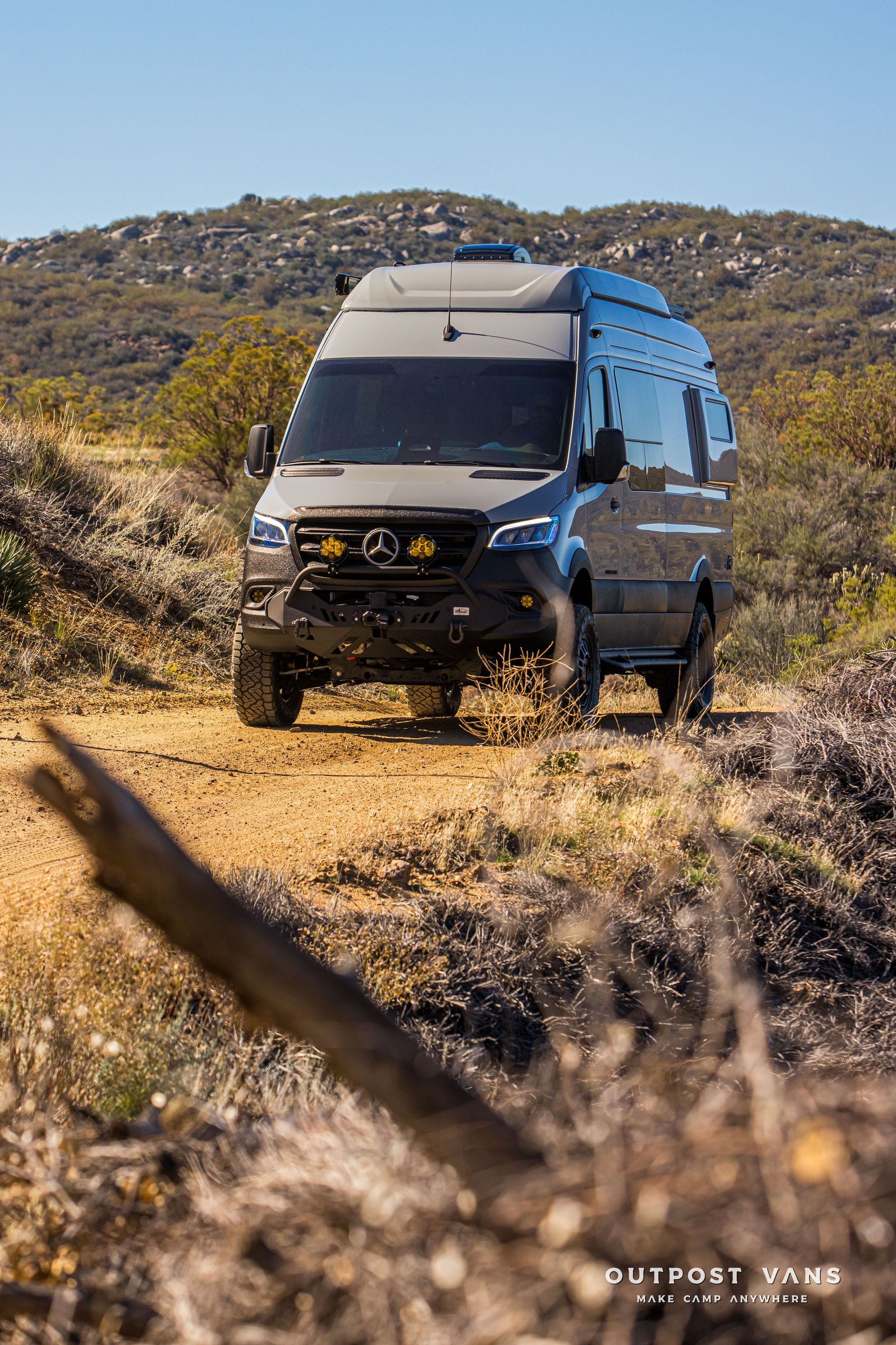Gray Mercedes Sprinter van driving on a dirt road in a rugged landscape with vegetation.