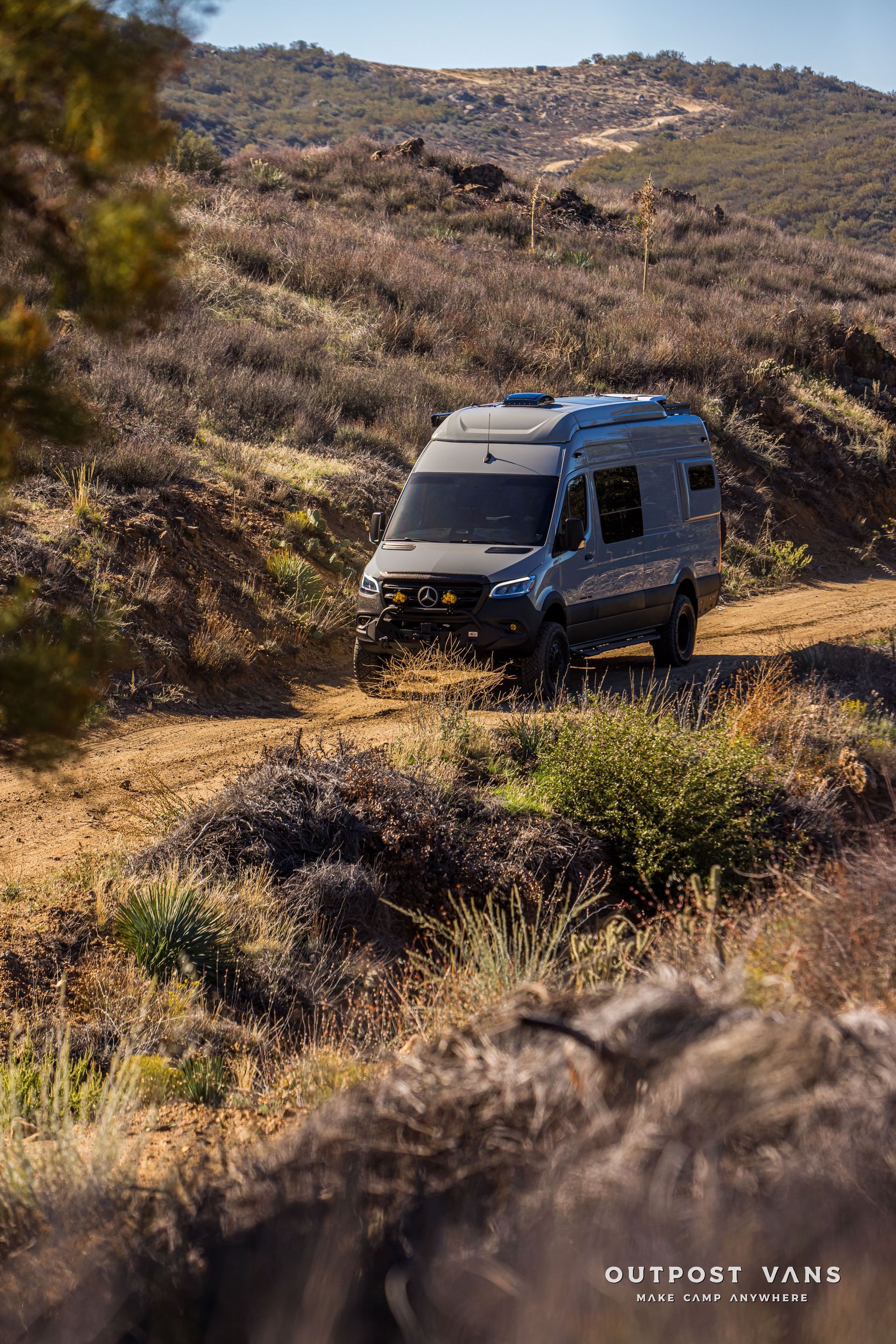 Gray camper van driving on a dirt road in a sunny, dry landscape.