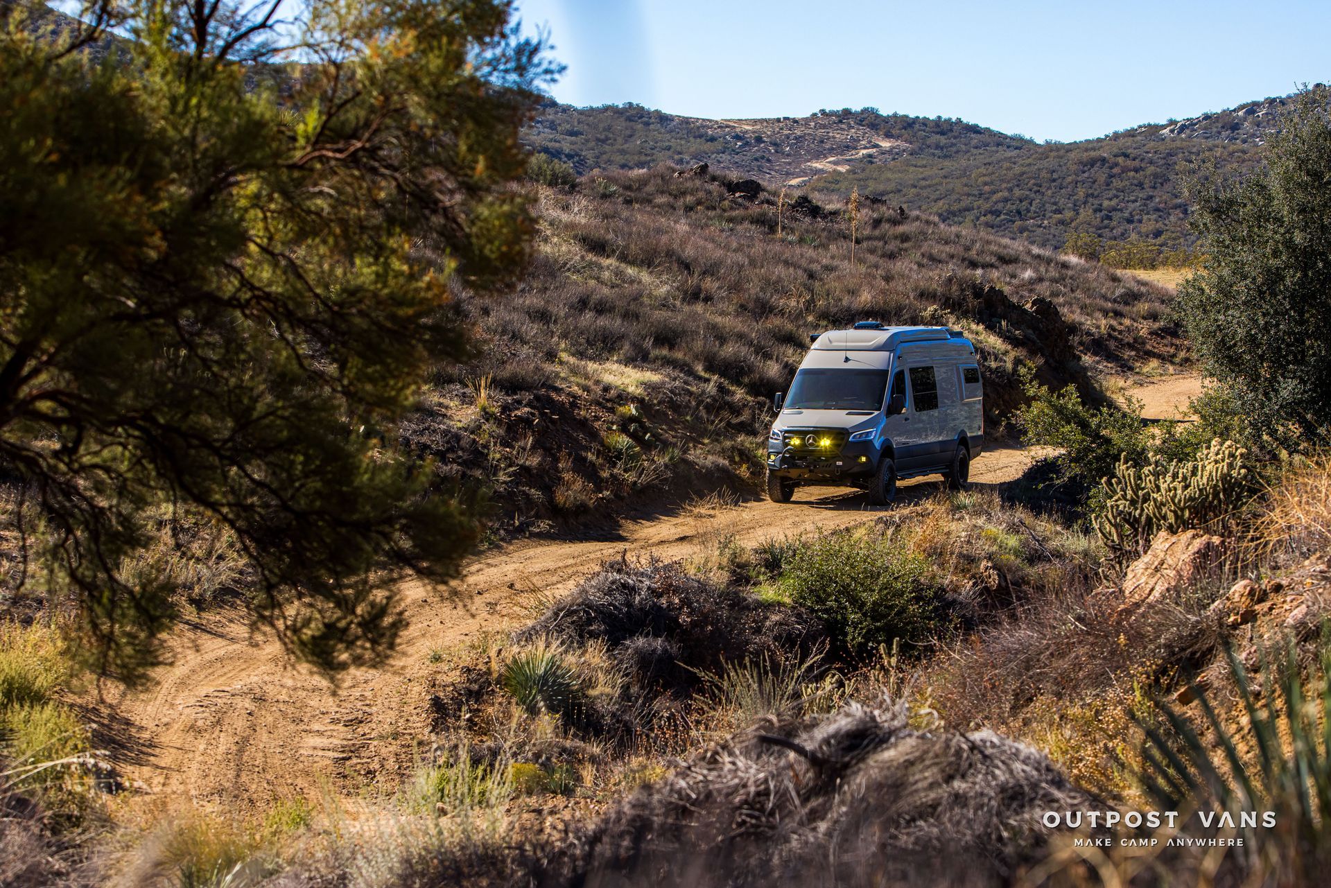 A gray van travels on a dirt road through a hilly, arid landscape.