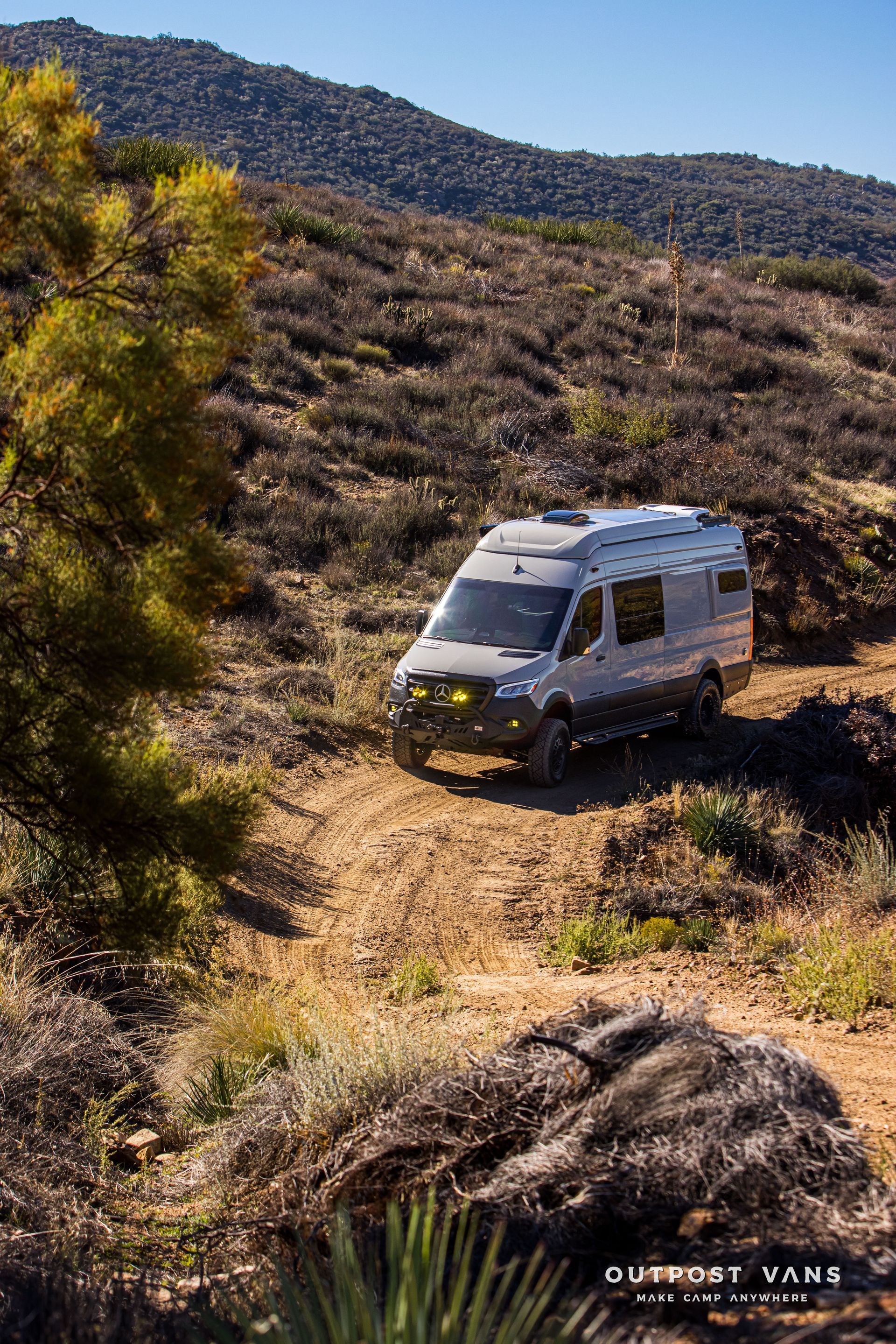 Gray camper van driving on a dirt road in a hilly, arid landscape under a clear blue sky.