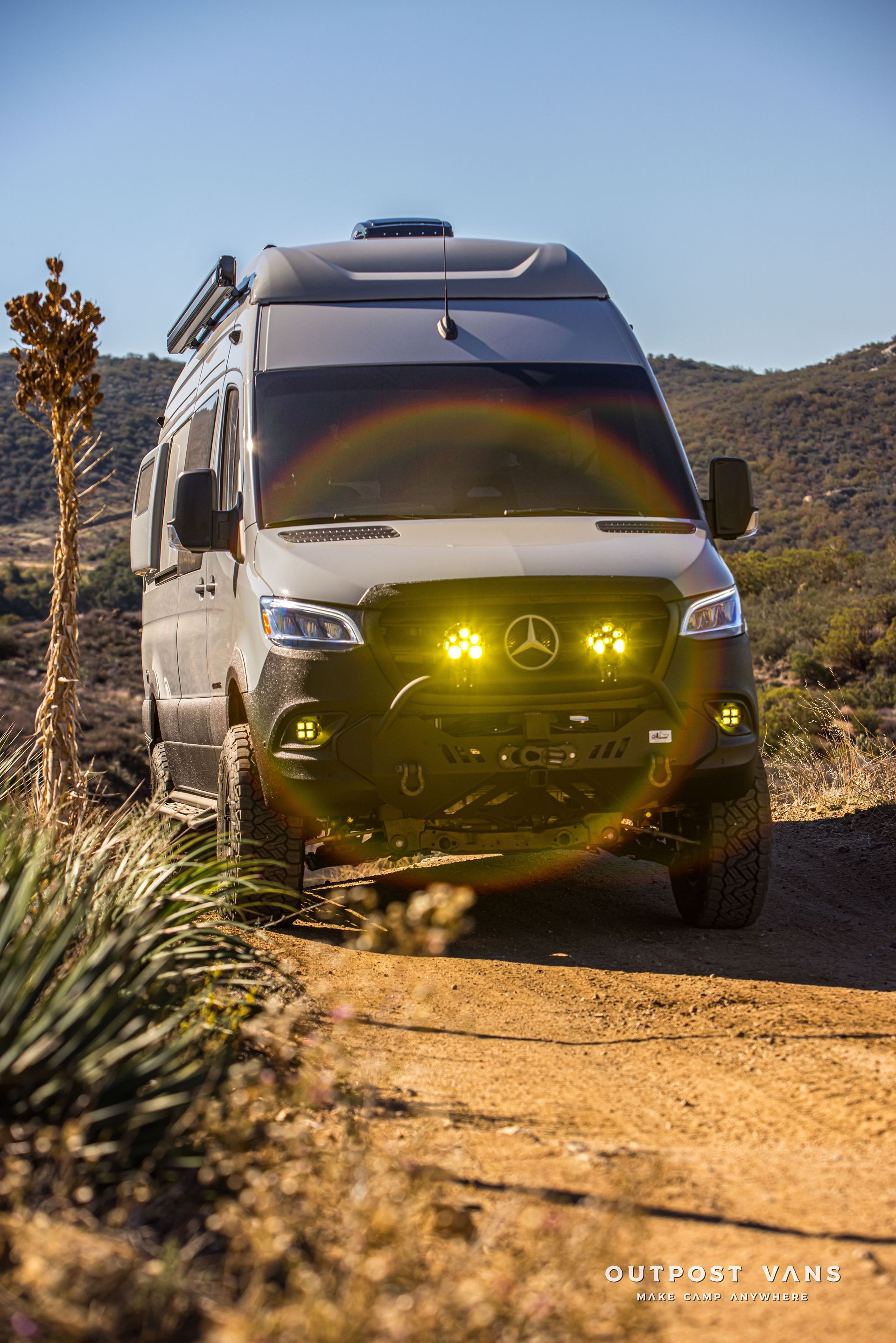 Gray Mercedes Sprinter van, off-road, with yellow lights, on a dirt road in a desert landscape.