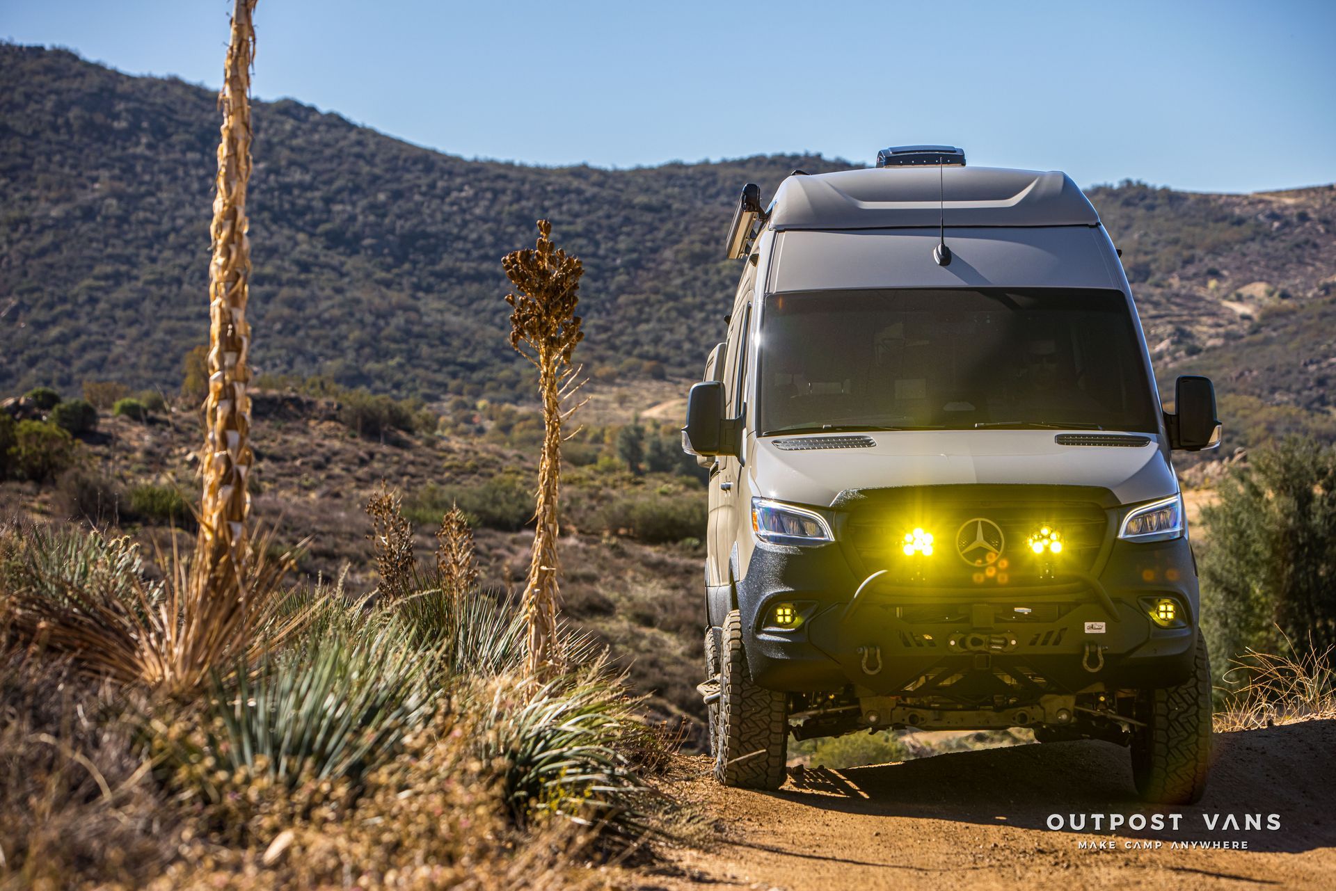 Off-road gray van with yellow fog lights on a dirt road in a dry, mountainous landscape.