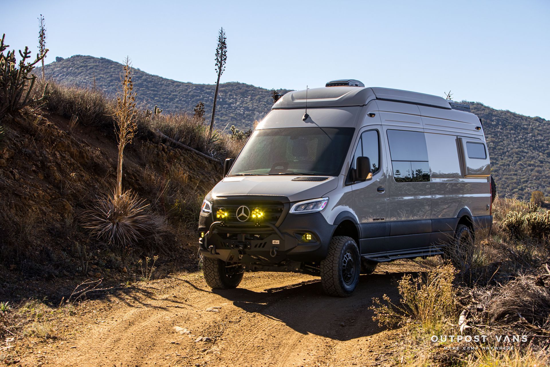 Gray off-road van on a dirt road, with yellow headlights and a desert backdrop.