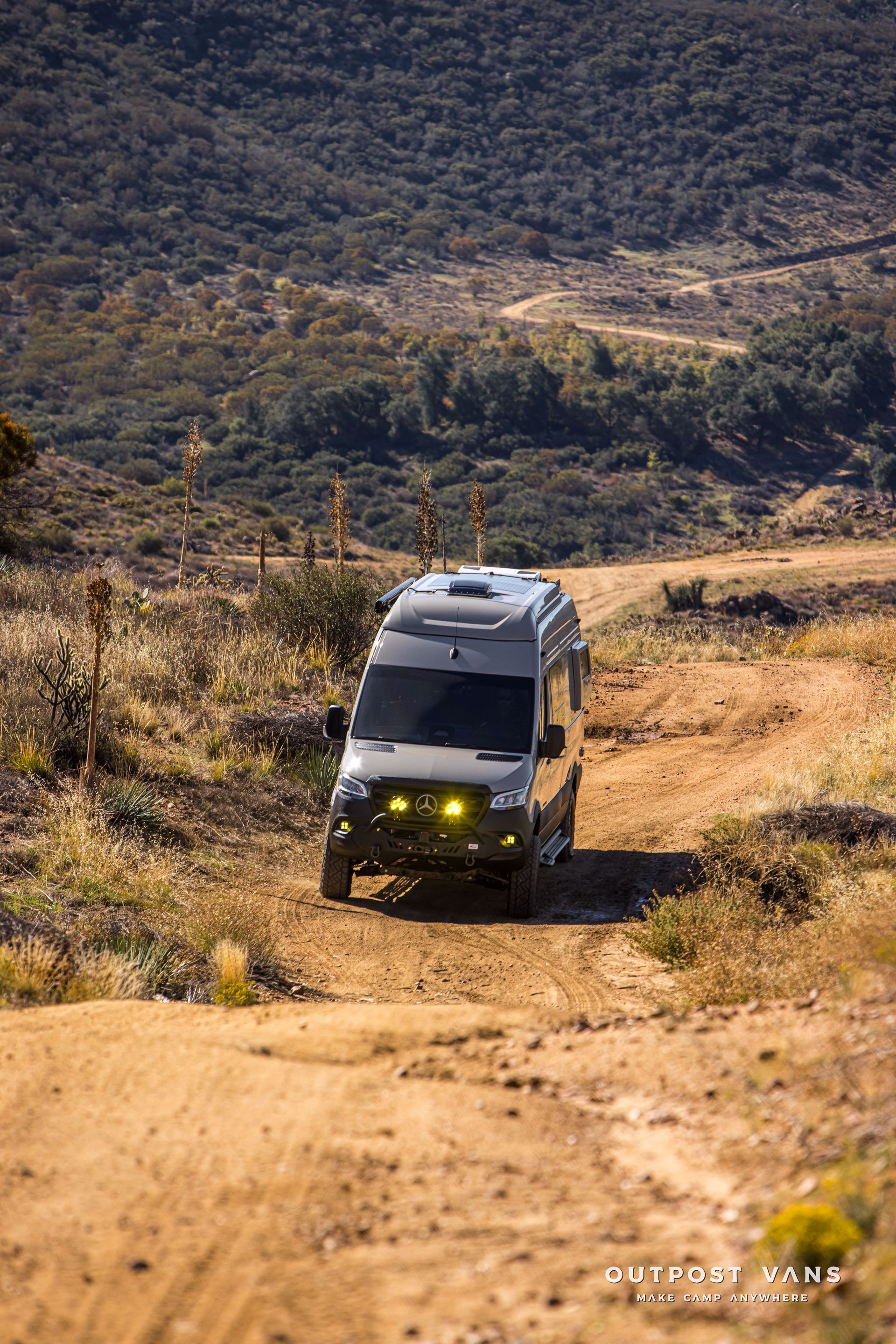 Off-road van drives up dirt road on a sunny day in a hilly, dry landscape.