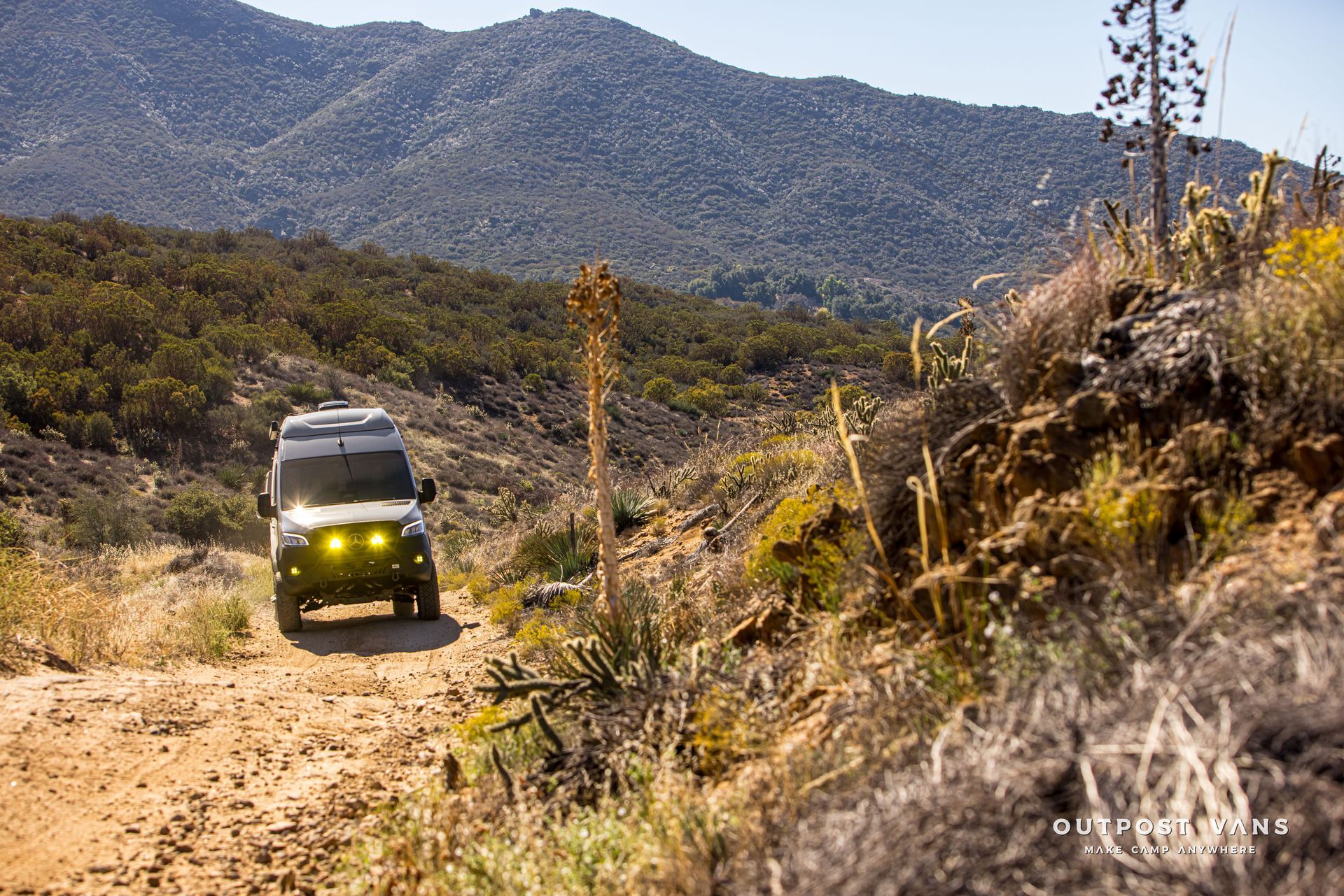 A camper van driving on a dirt road in a mountainous, sunny landscape.