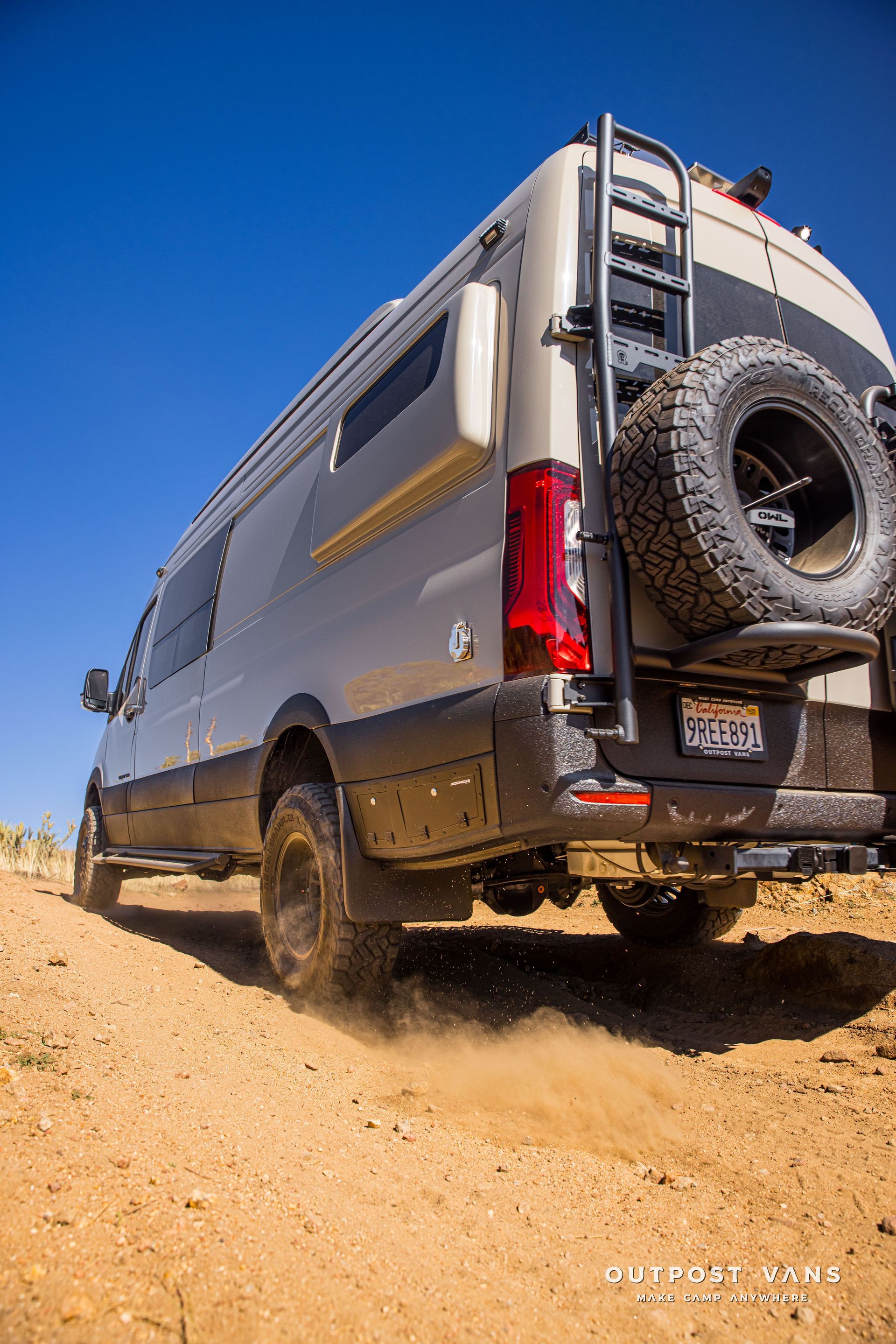 Off-road adventure van on a dirt road, kicking up dust. Tan and black vehicle with spare tire and ladder.