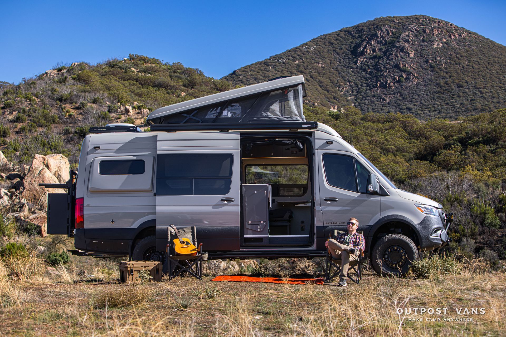 Gray camper van with open door and pop-up roof in a mountain setting. A person sits nearby.