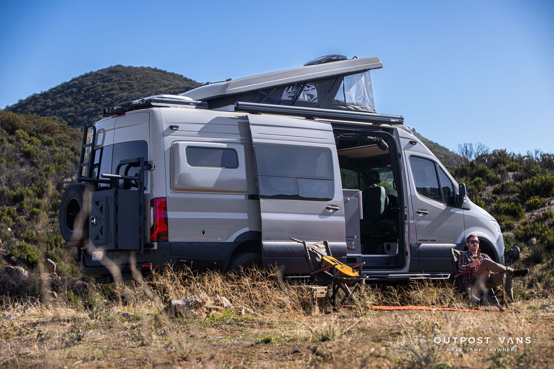 Silver camper van with pop-up roof parked in a field, door open. A person sits nearby in a folding chair.