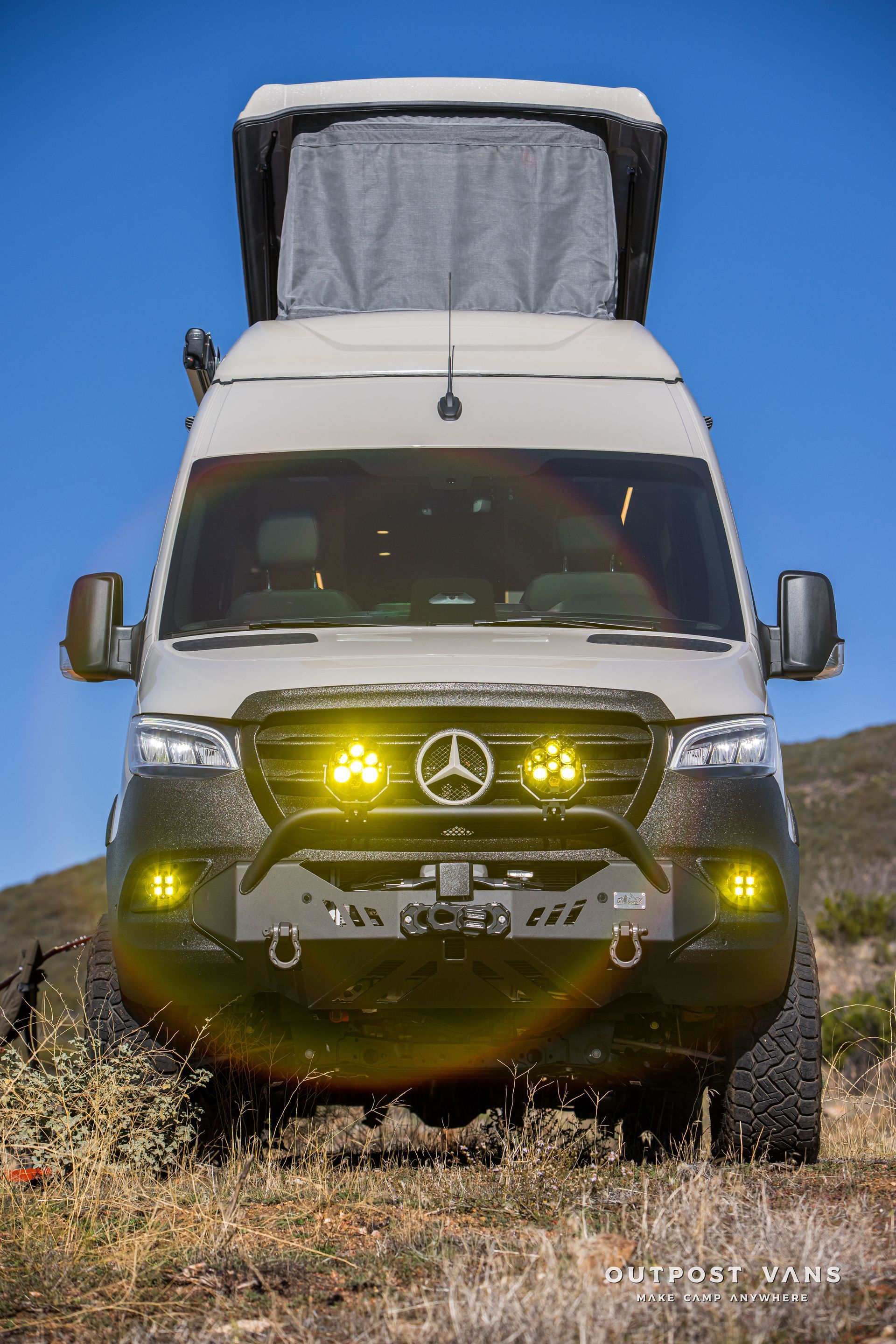 Off-road modified gray Mercedes Sprinter van with pop-up roof and yellow lights parked on a dirt path under a blue sky.