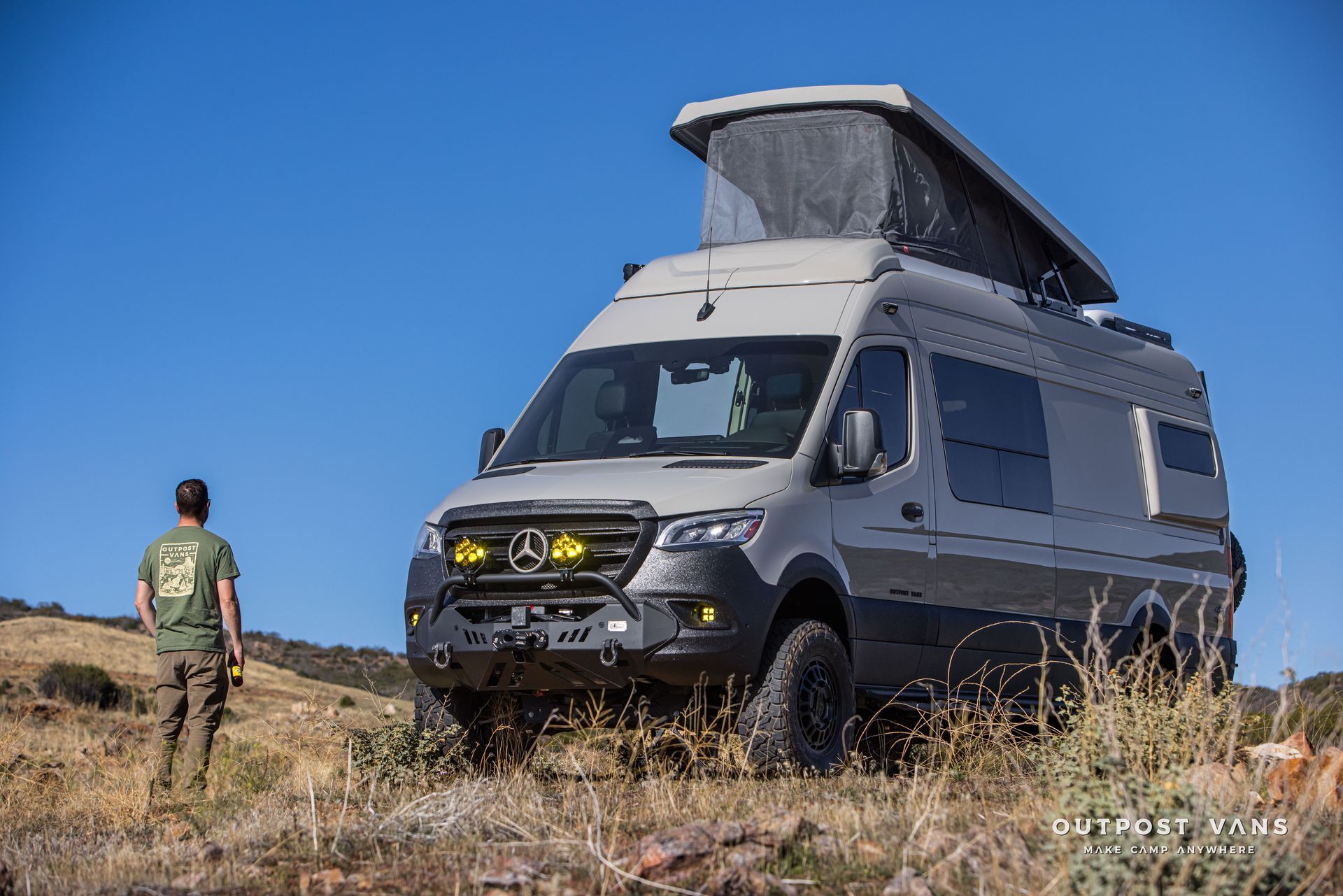 Gray camper van with a pop-up roof parked outdoors. A person stands nearby, looking at it.