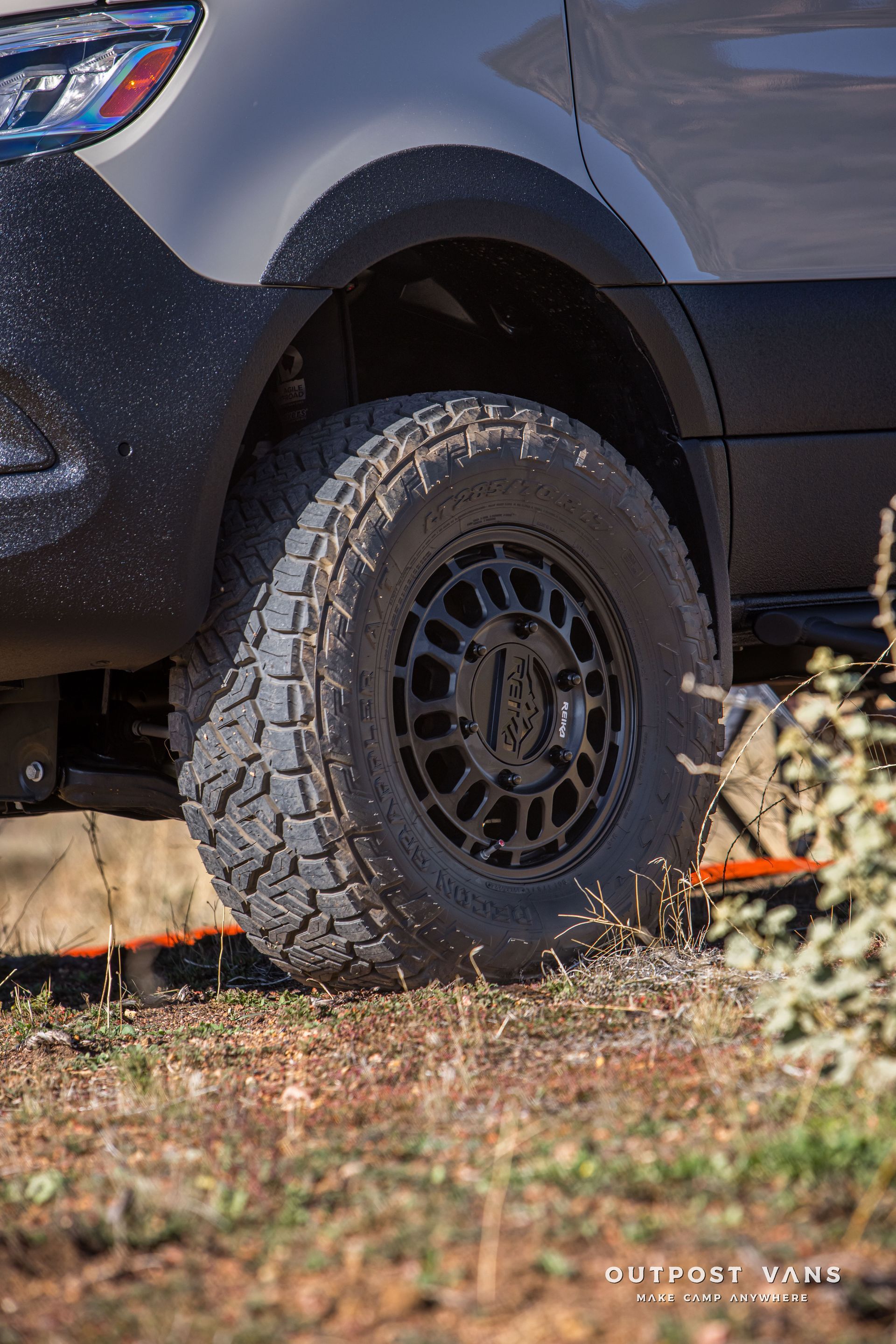 Black van tire on a dirt road, spraying gravel. Black wheel, light gray van body.