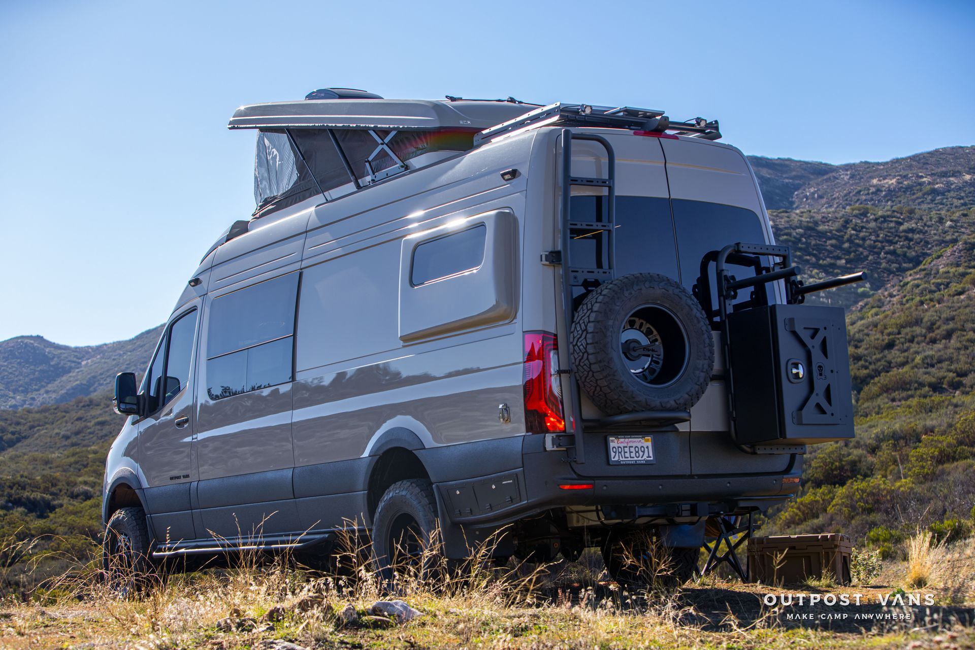 Tan and gray camper van with pop-up roof and spare tire carrier parked on a hillside.