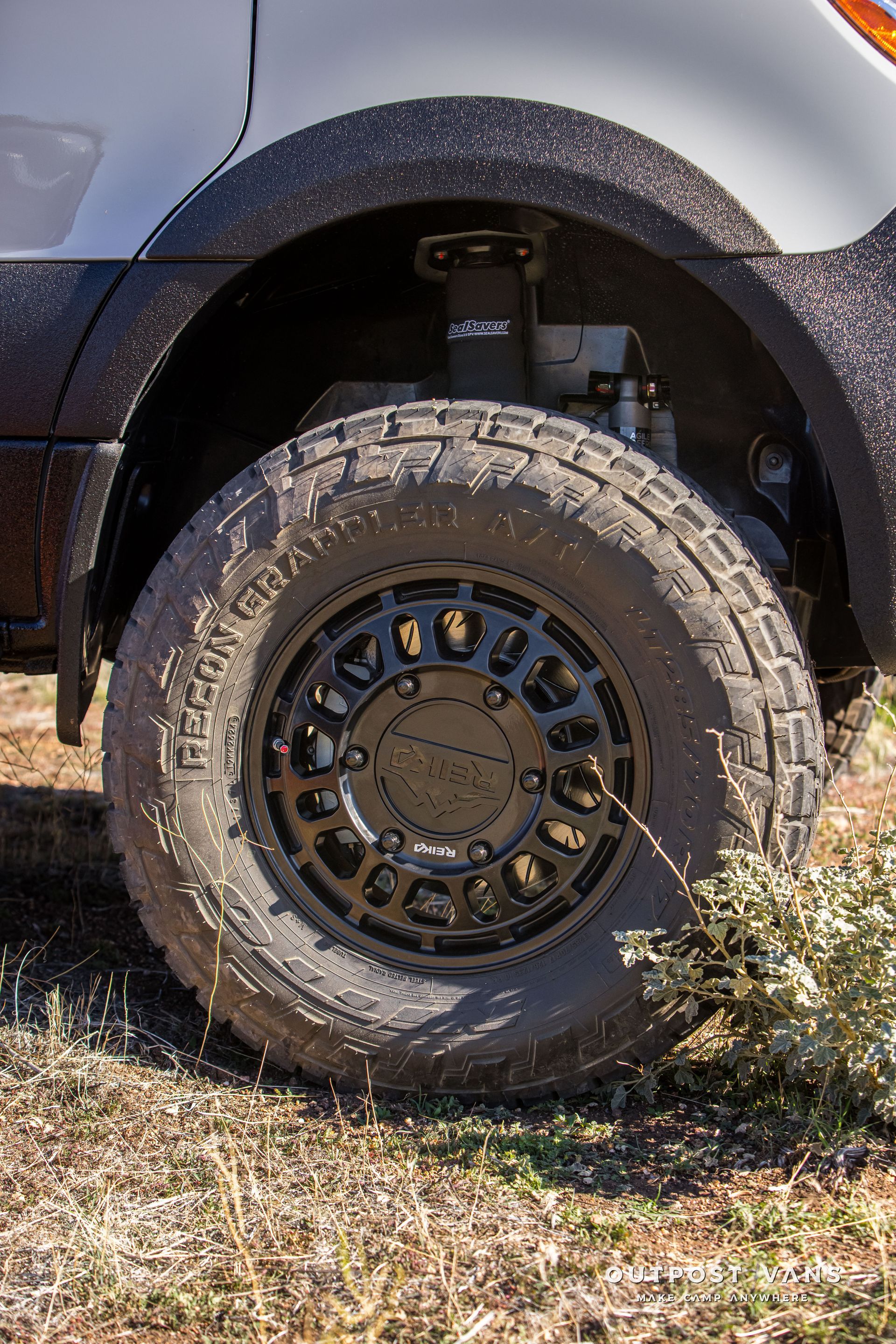 Black off-road tire on a black wheel under the fender of a white van with black trim; parked on dirt.