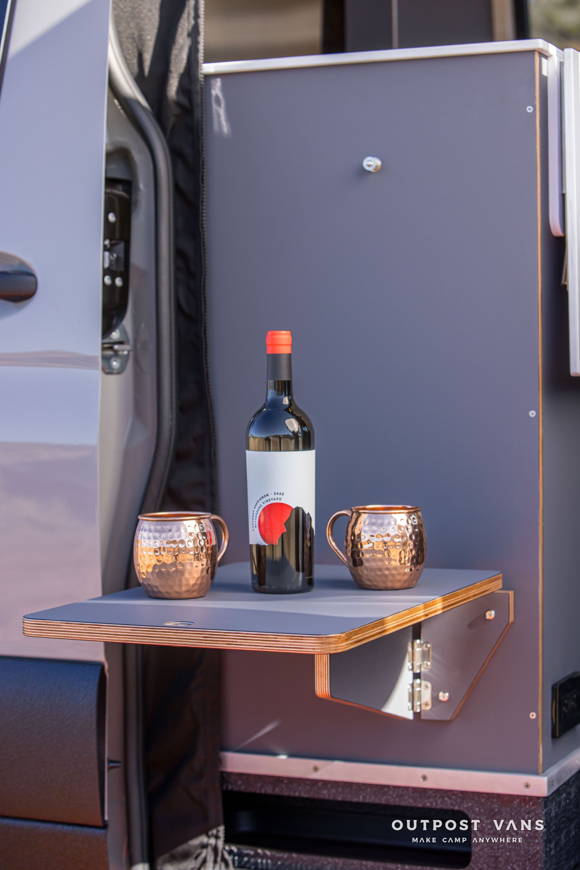 Wine bottle and copper mugs on a fold-down table attached to the side of a campervan.