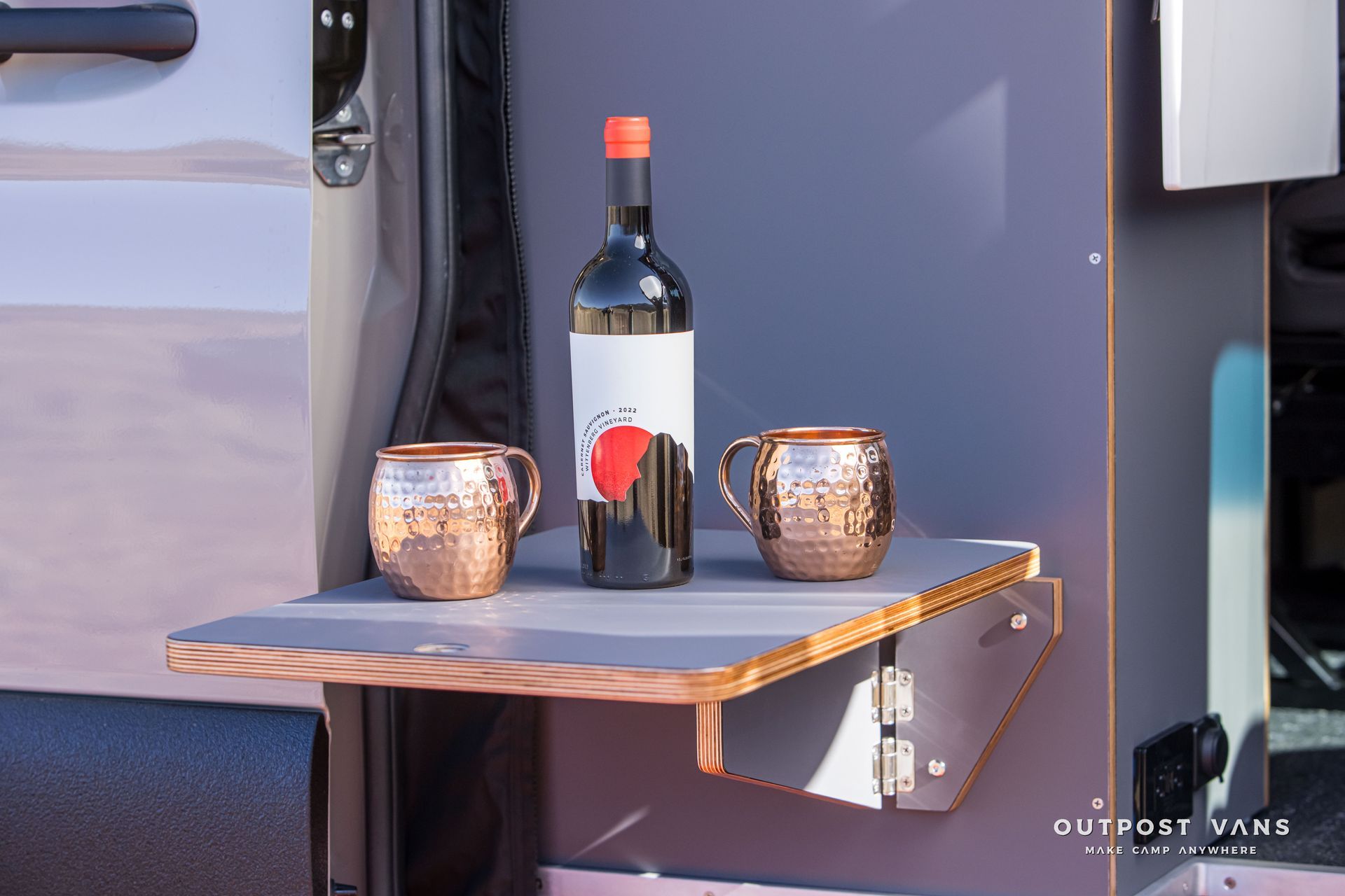 Fold-down shelf with copper mugs and wine bottle in front of a van interior.