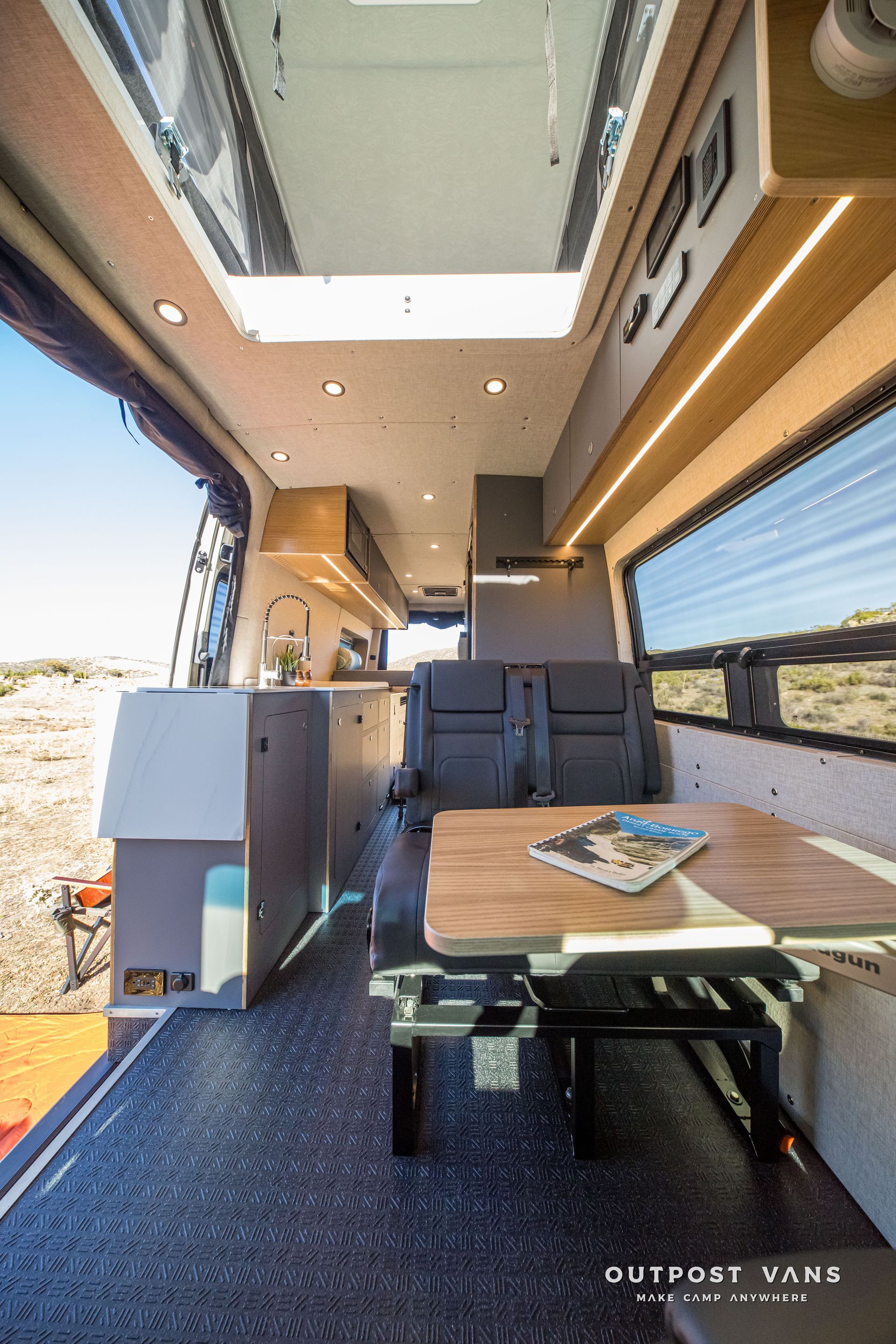 Interior view of a camper van with wood accents, a table, and windows overlooking a desert landscape.