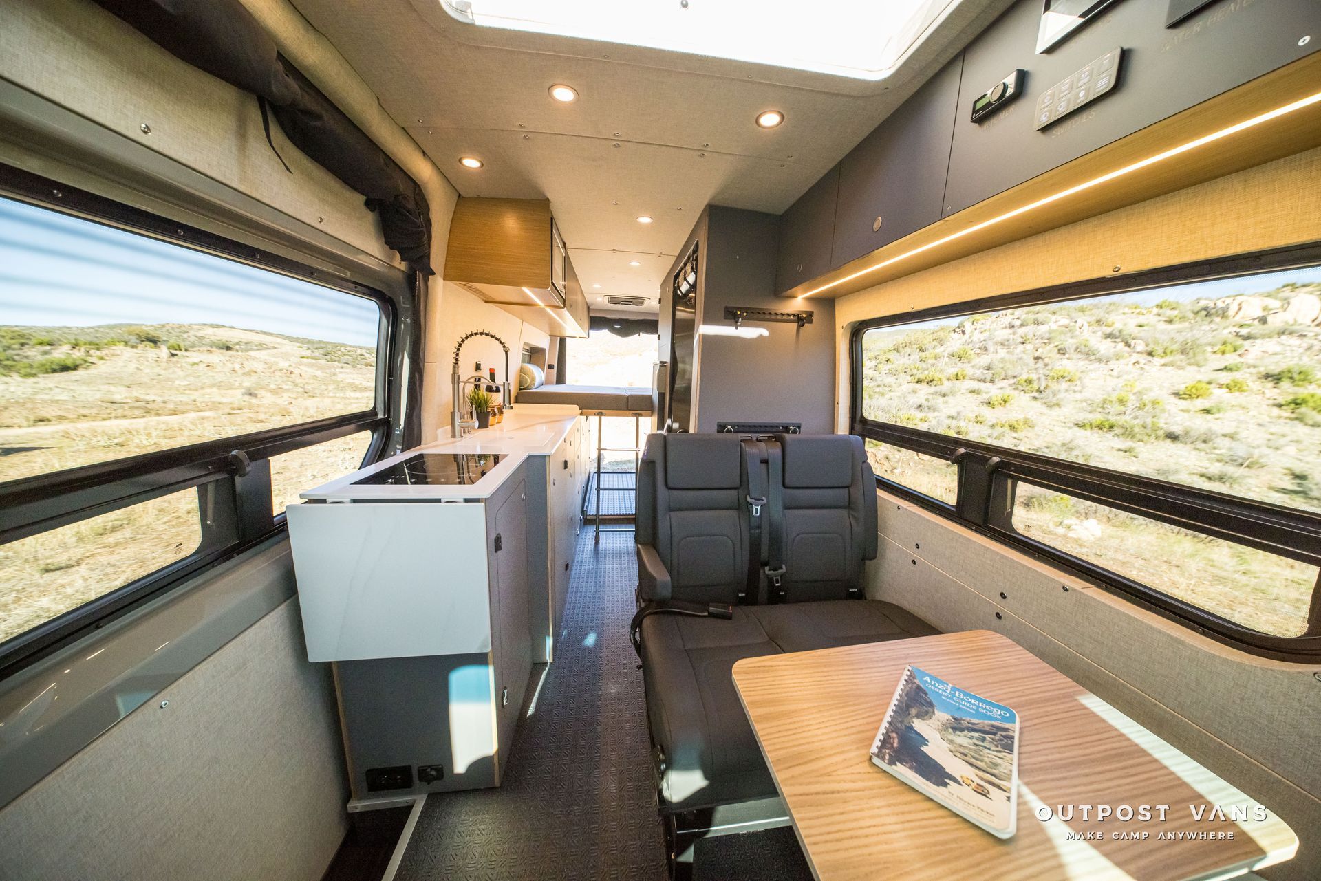 Interior view of a campervan with a kitchen, seating area, and windows overlooking a landscape.