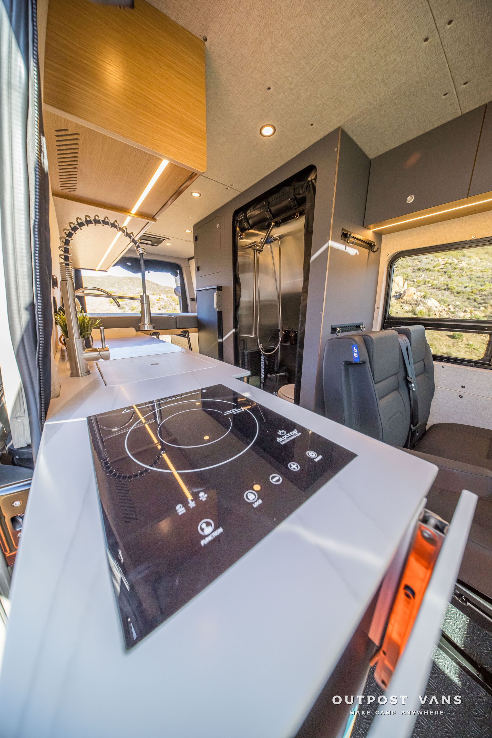 Van interior with modern kitchen, cooktop, shower, and seating area. White countertops, gray walls, natural light.
