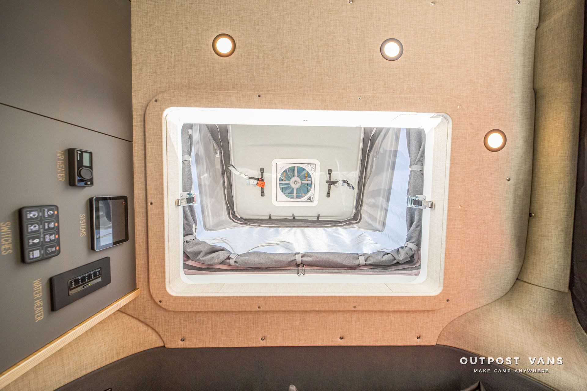 Interior of camper van: overhead view through a window with a fan, control panels on the wall, neutral tones.