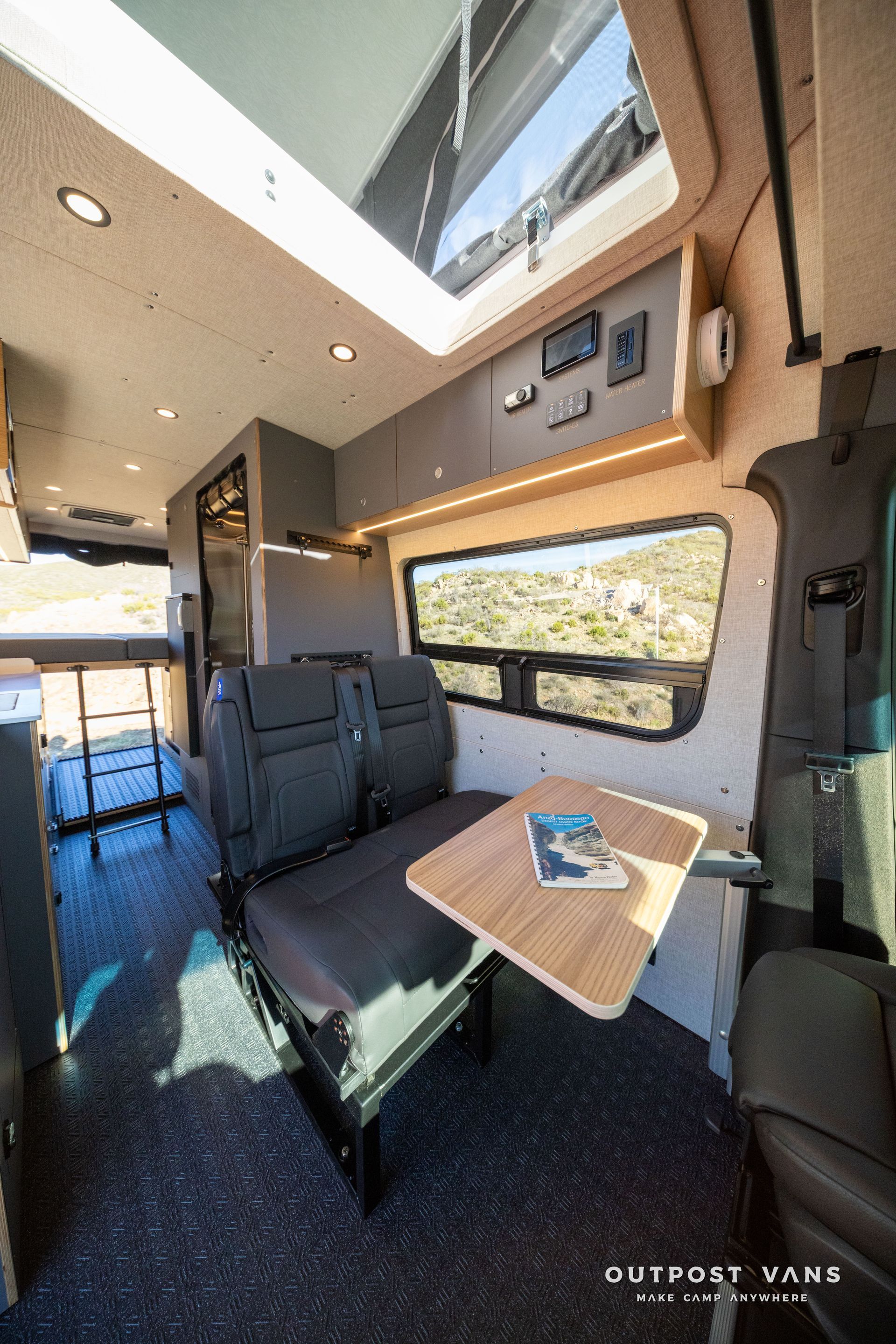Interior of a camper van with seating, a table, and overhead storage; natural light streams in.