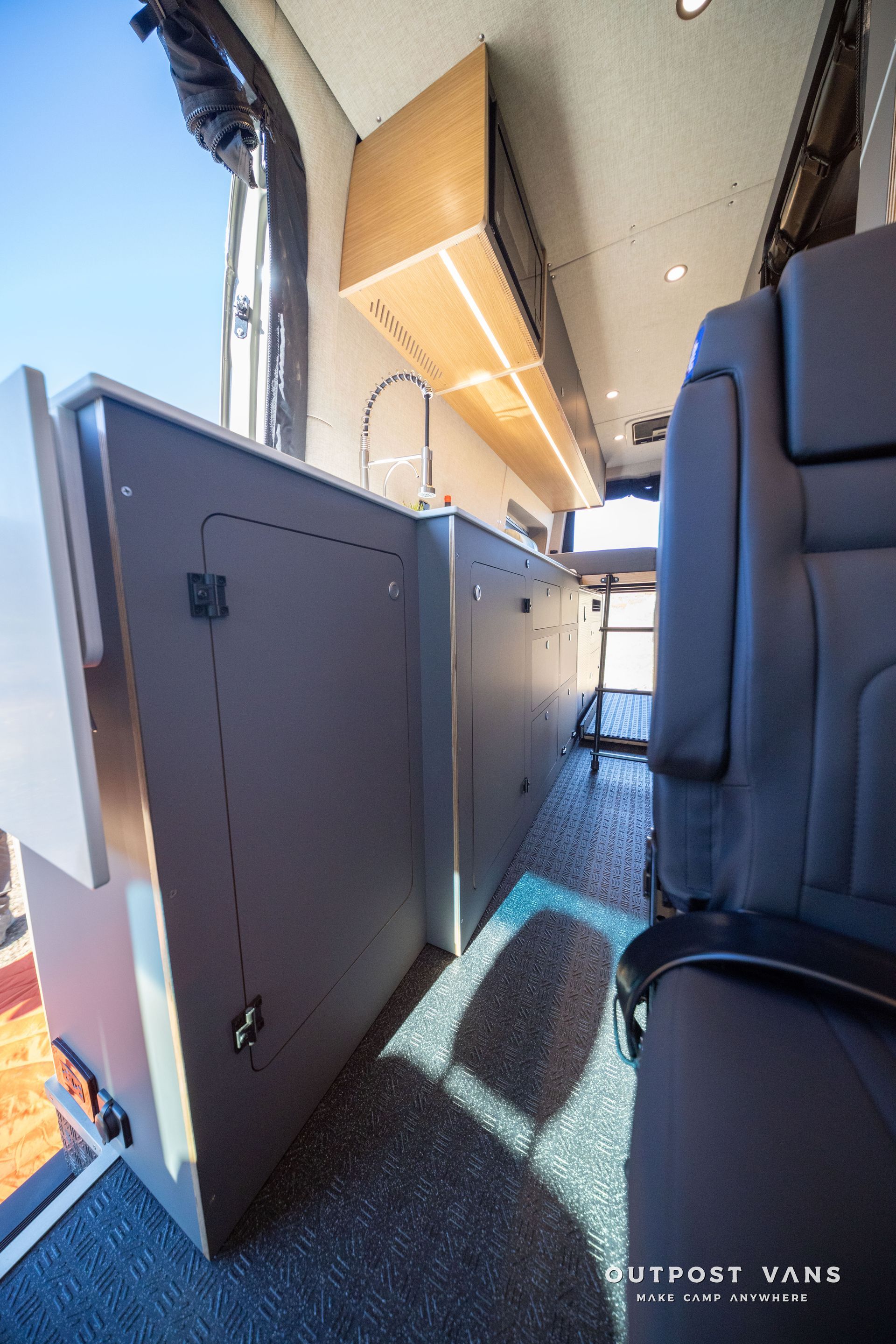 Interior of a camper van with cabinets, a seat, and a window. Gray cabinets and blue flooring are visible.