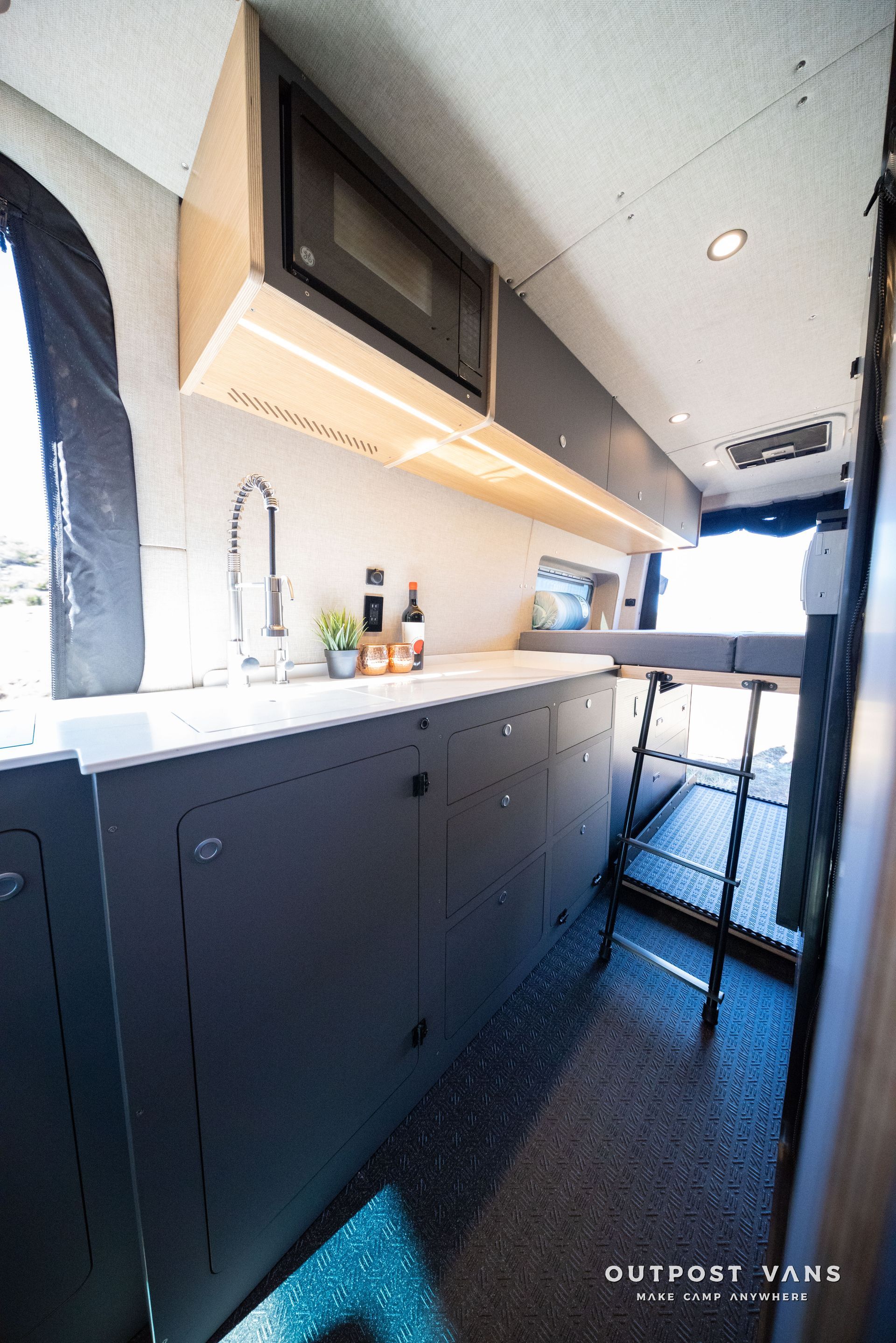 Interior of a van with a kitchen area. Dark gray cabinets, white countertop, microwave, and sink.