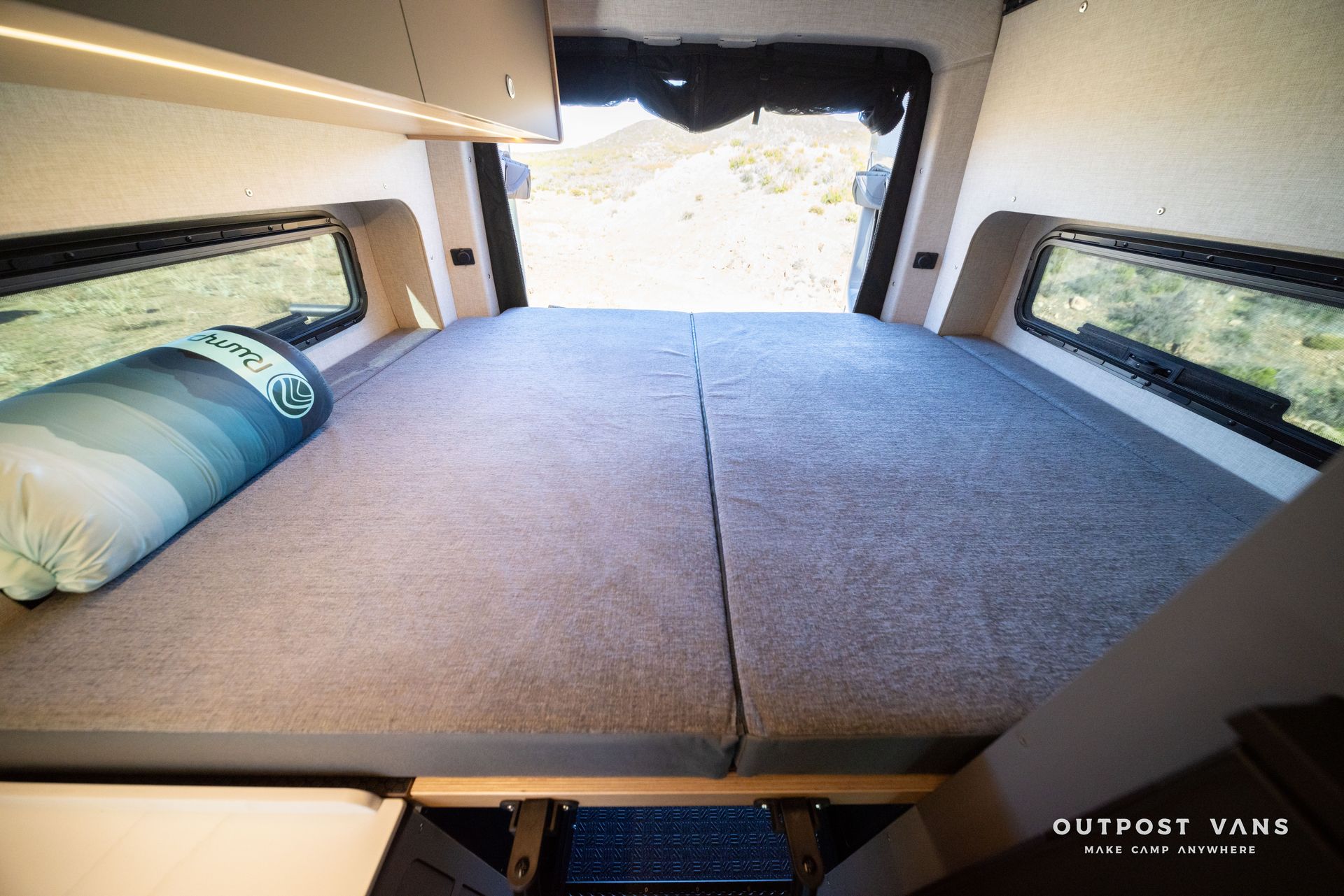 Interior of a camper van with a bed, windows, and open doors revealing an outdoor view.
