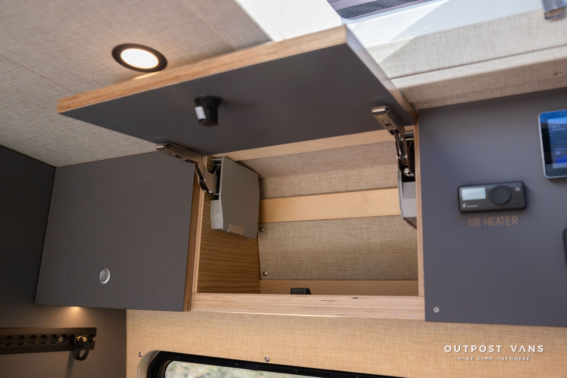 Gray overhead cabinet in a van interior, open. Wood and gray tones. Recessed light.