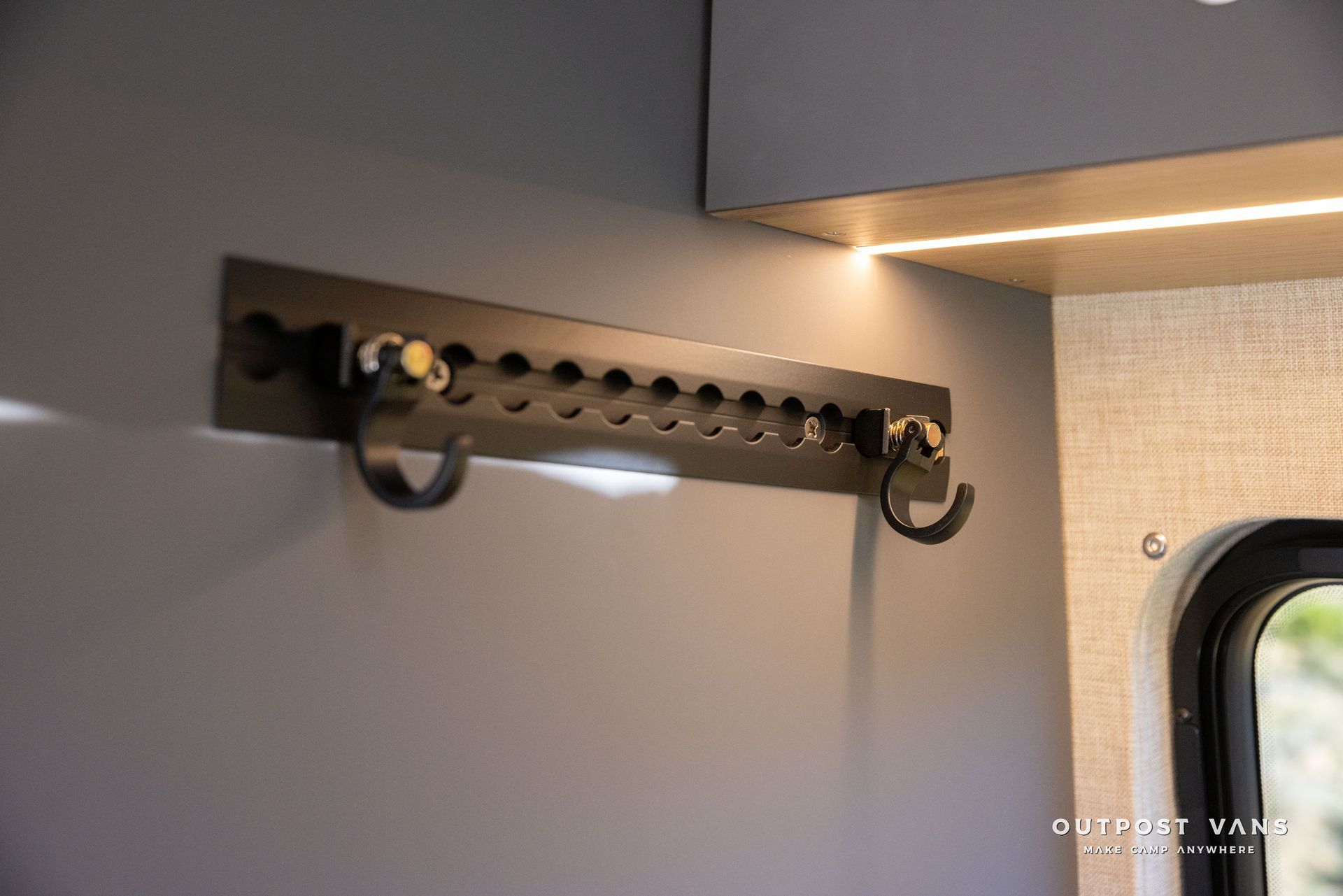 Dark metal coat rack with hooks mounted on a gray wall, lit by a strip light.