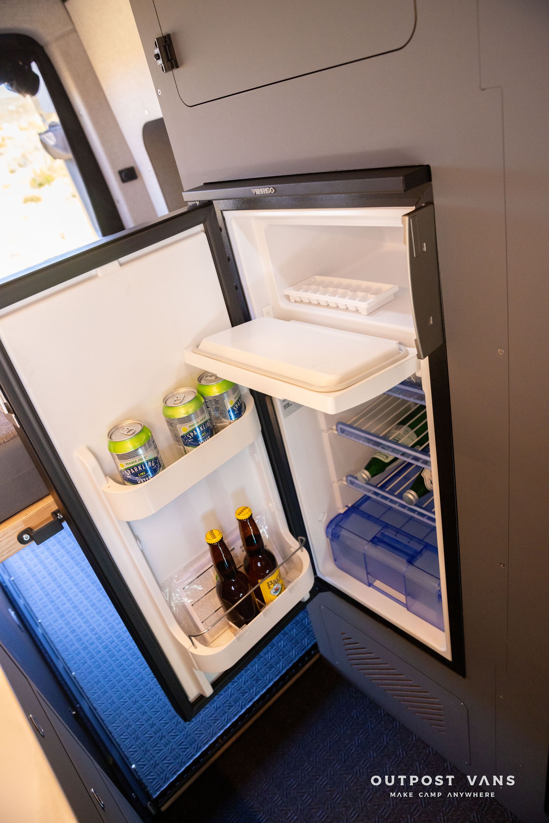 Open refrigerator in a van, stocked with drinks and food. Blue accents on the floor and seat.
