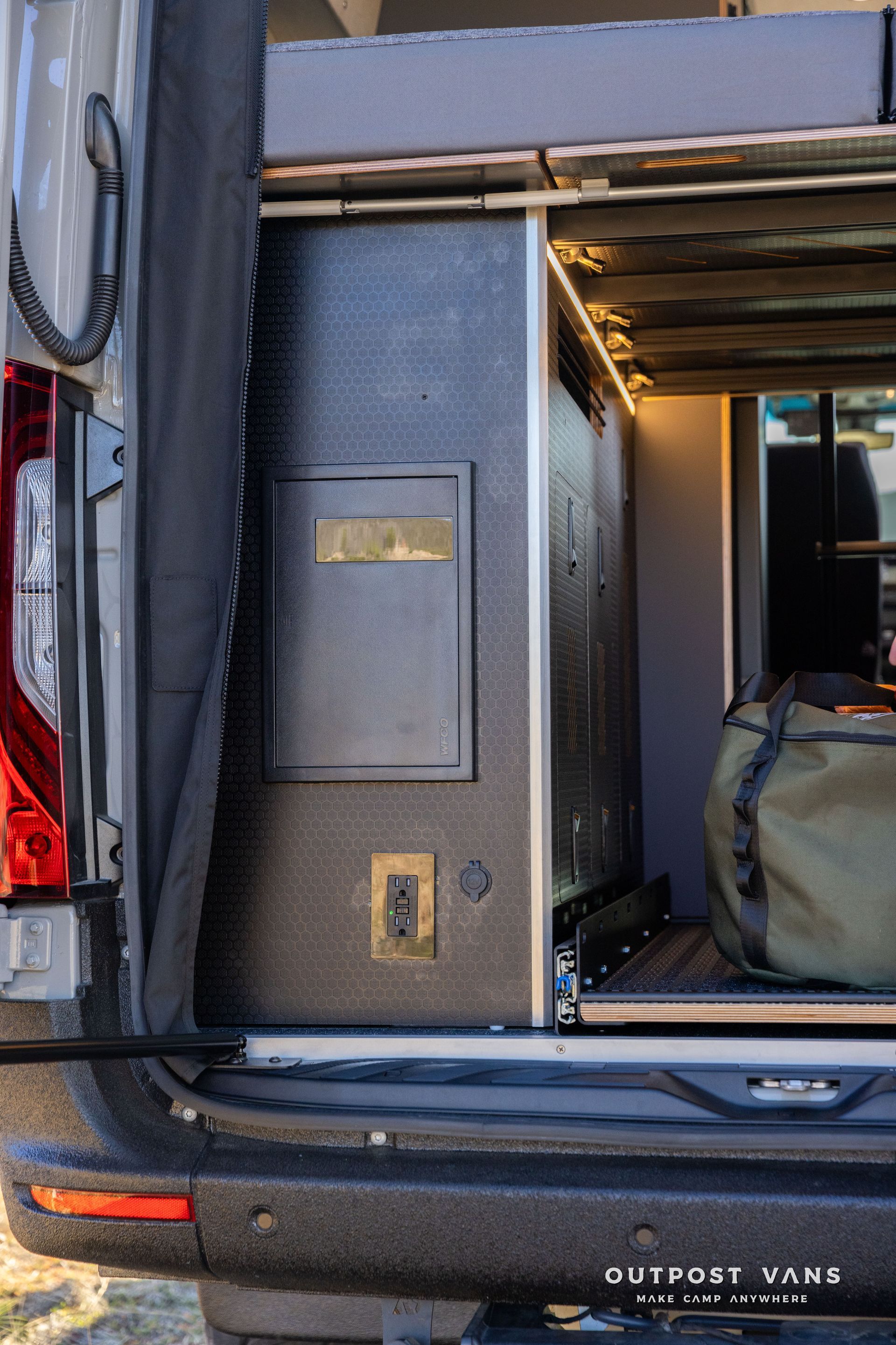 Close-up of a van's open rear compartment. Interior cabinet with outlet, drawer, and lights visible. Dark textured panels.
