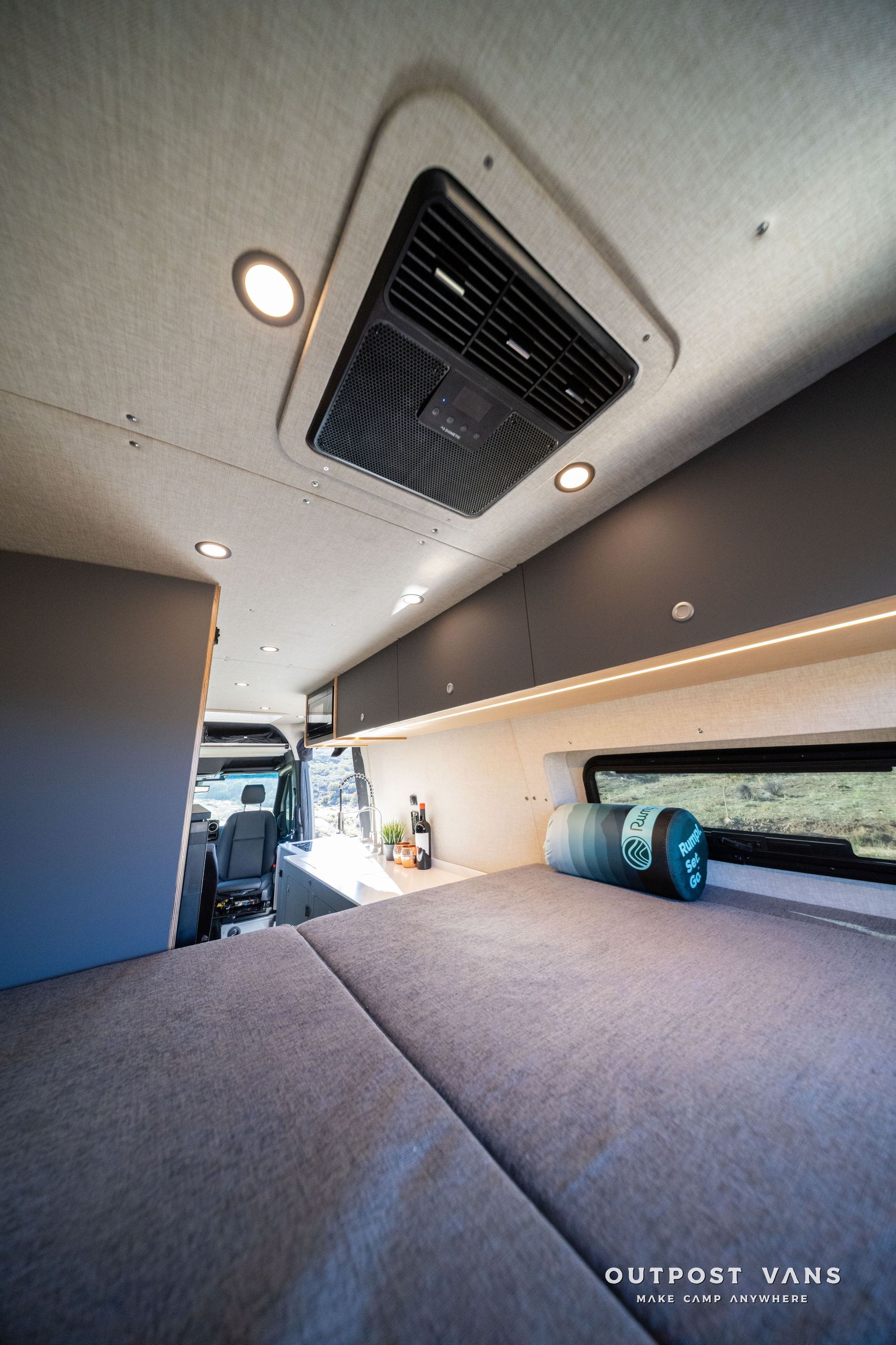 Interior of a camper van with bed, overhead storage, air vent, and lighting.