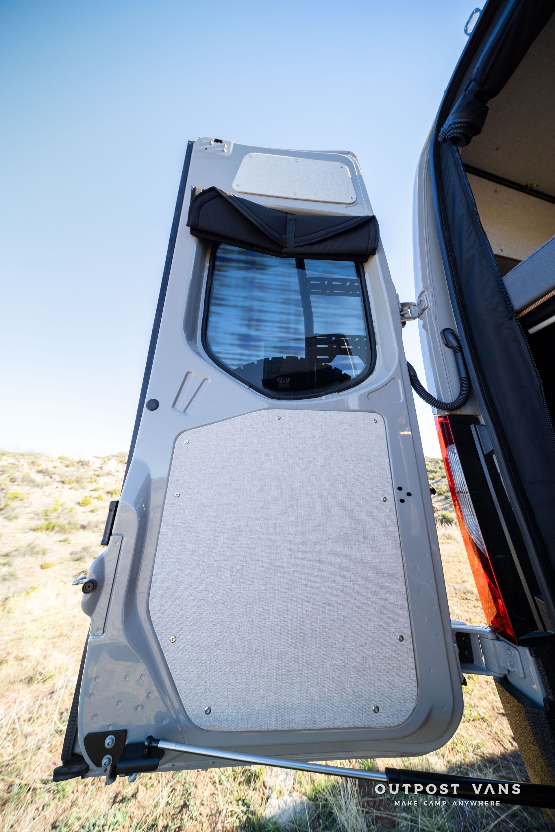Open grey van door, revealing window with a shade, in a field, sunny day.