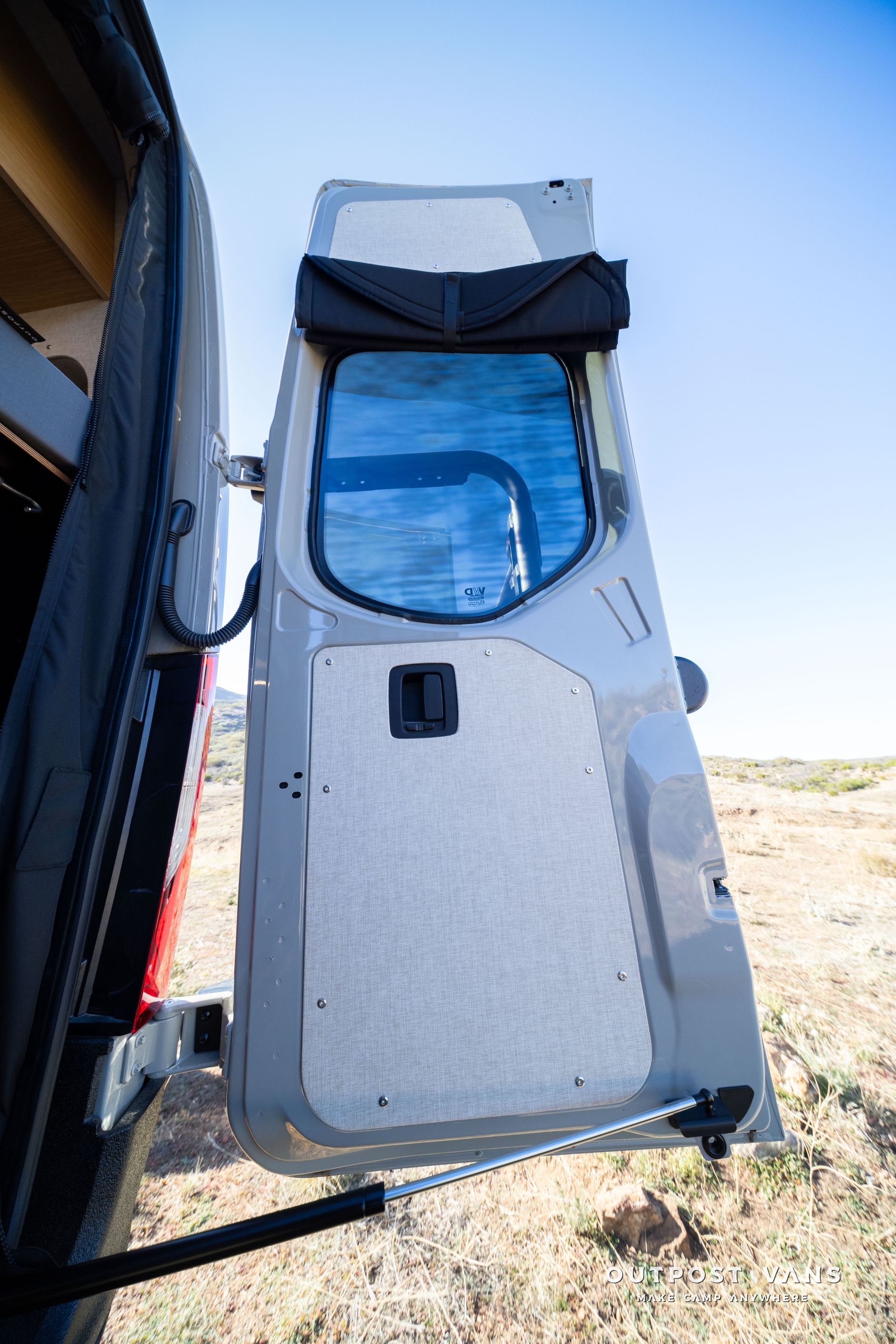 Open van door with gray paneling, blue window, and black awning in a sunny outdoor setting.