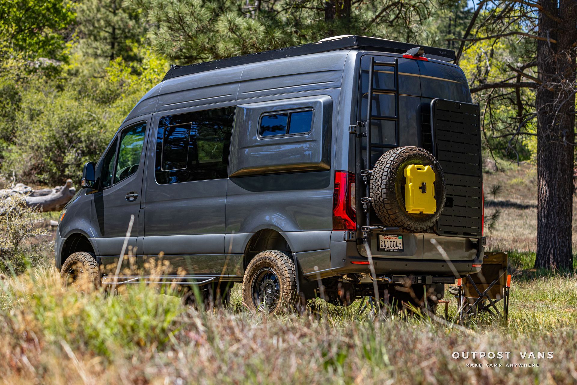 Gray camper van parked in a sunny forest clearing, viewed from the rear side.