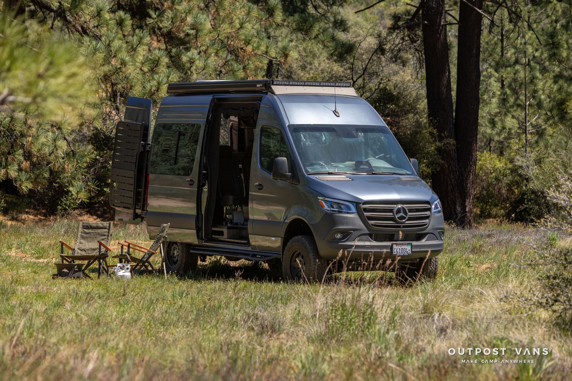 Gray camper van parked in a grassy woodland clearing with chairs outside