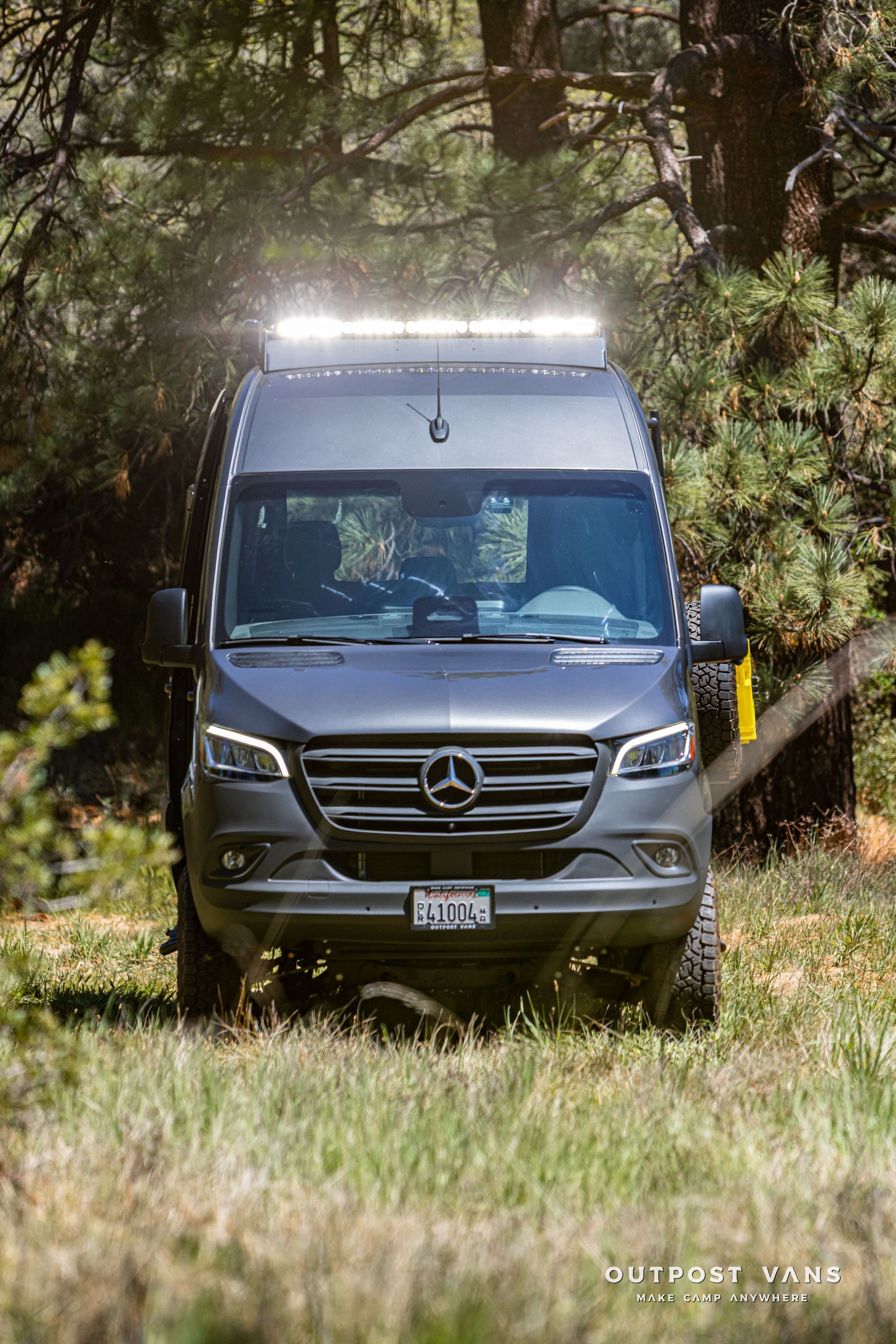 Gray Mercedes van driving on a grassy forest track with roof lights on