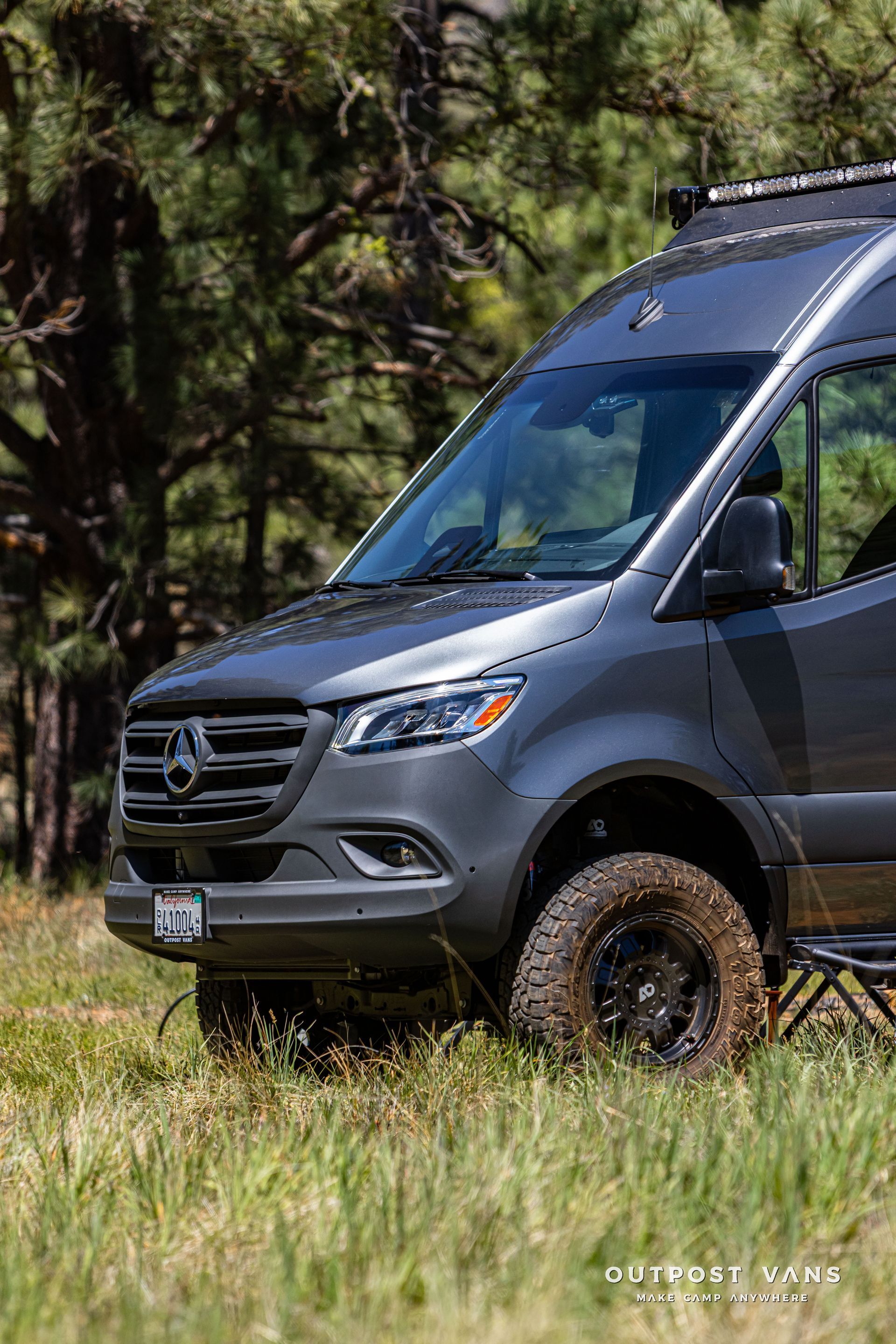 Gray van parked in grass with off-road tires in a wooded area