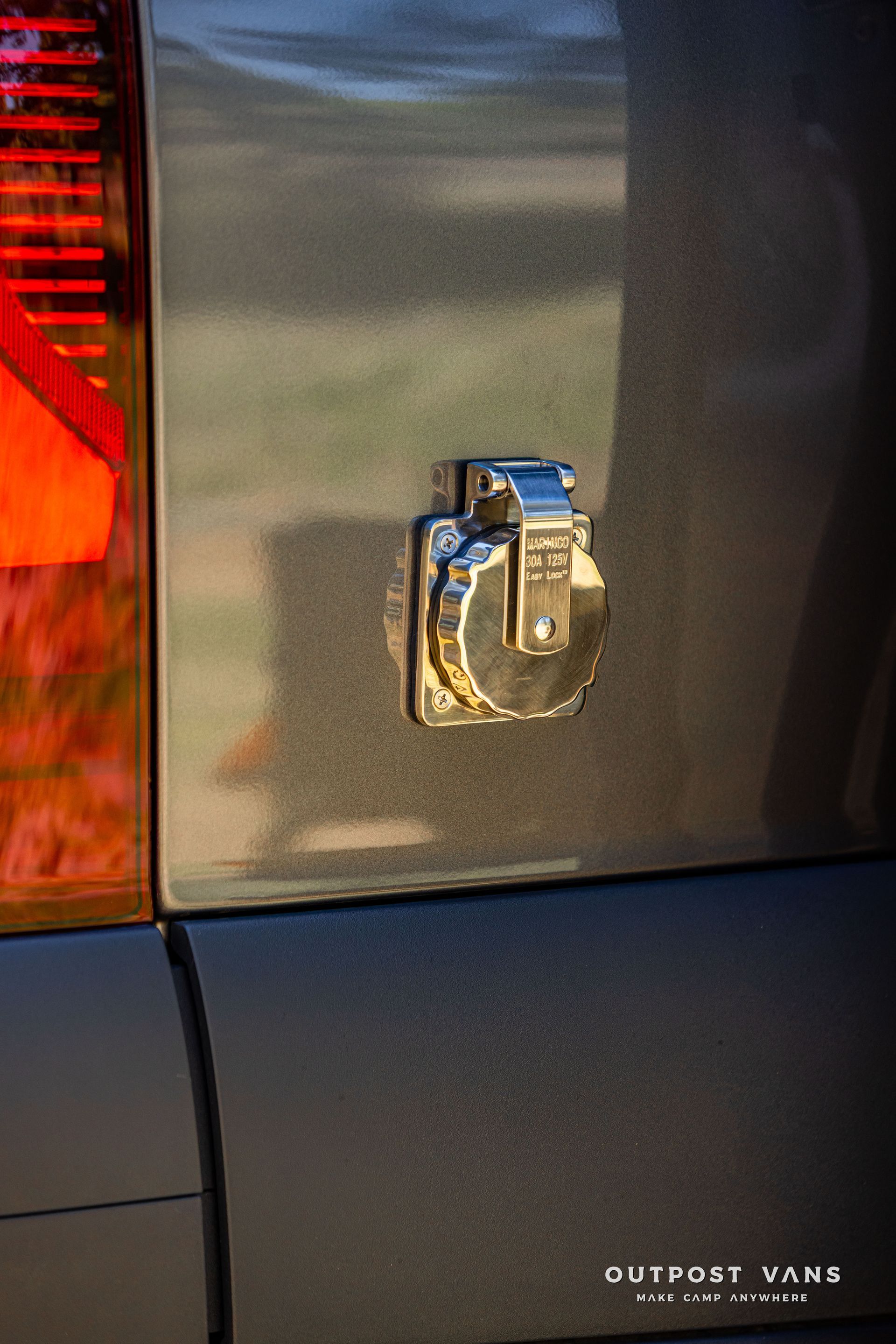 Close-up of a chrome car trunk latch on a dark vehicle, with a red taillight at left.