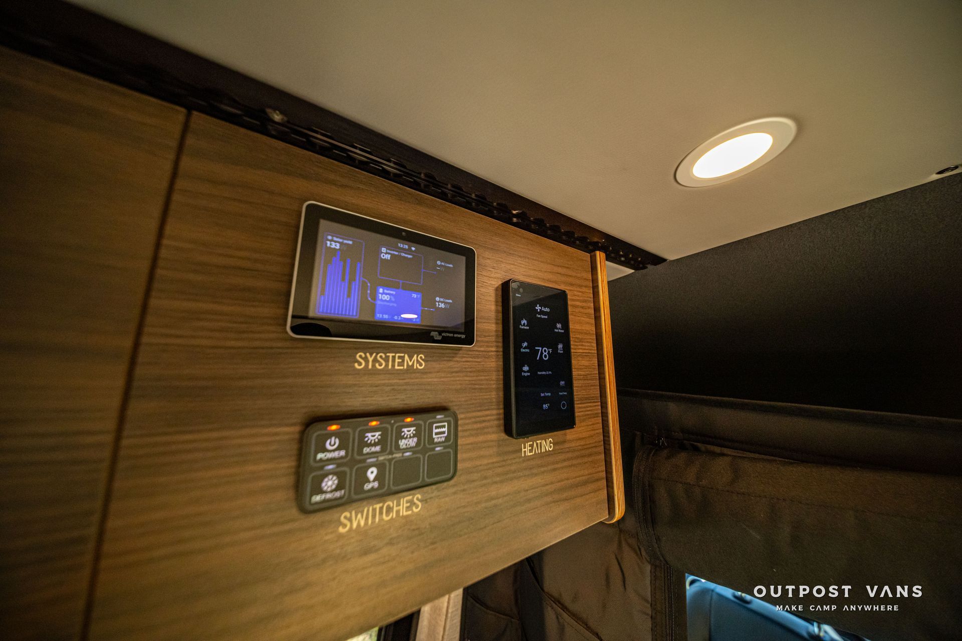 Wood-paneled RV control panel with touchscreen and switches above a bunk bed and window shades