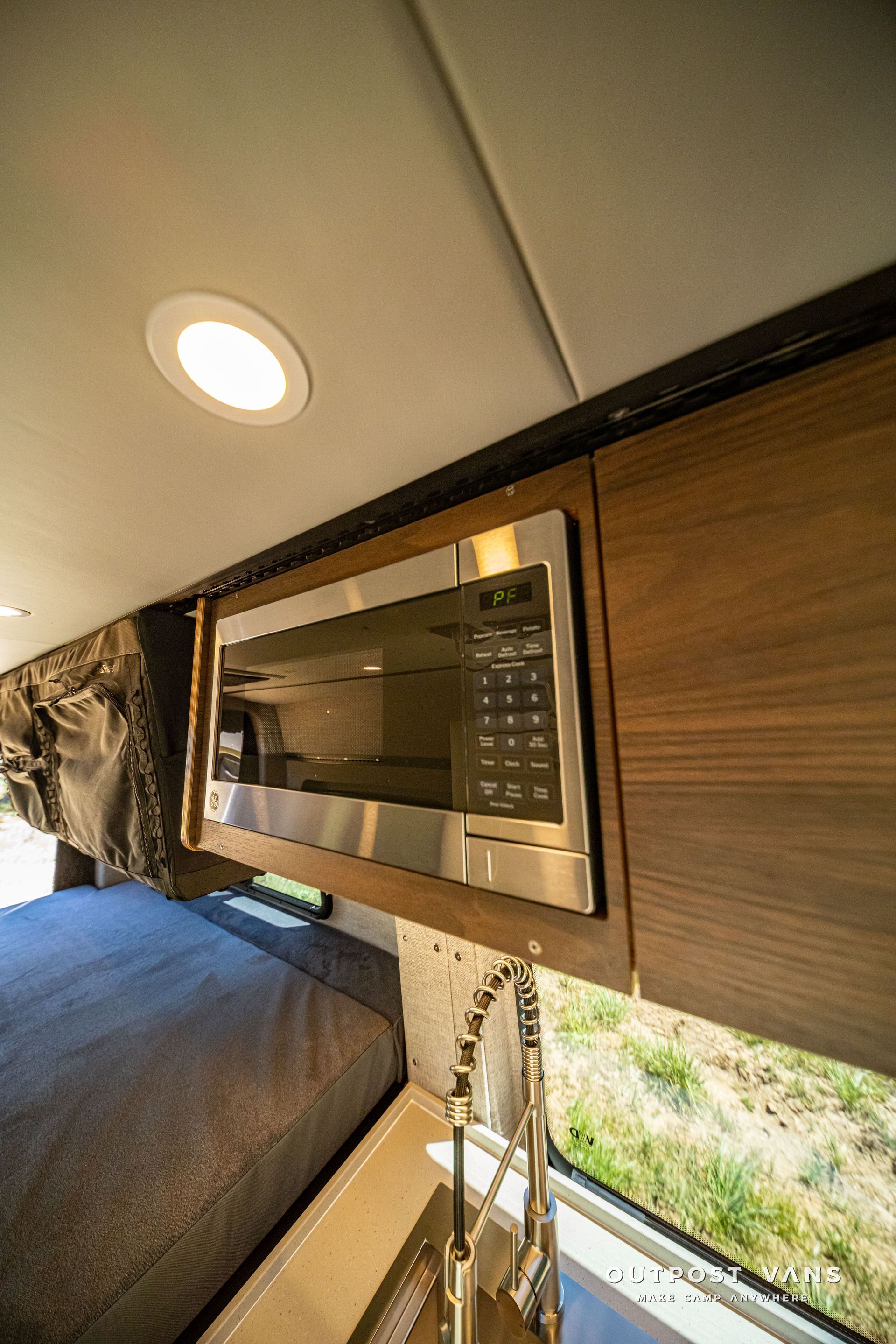 Wall-mounted microwave in a compact wood-paneled RV interior with a bed and window nearby