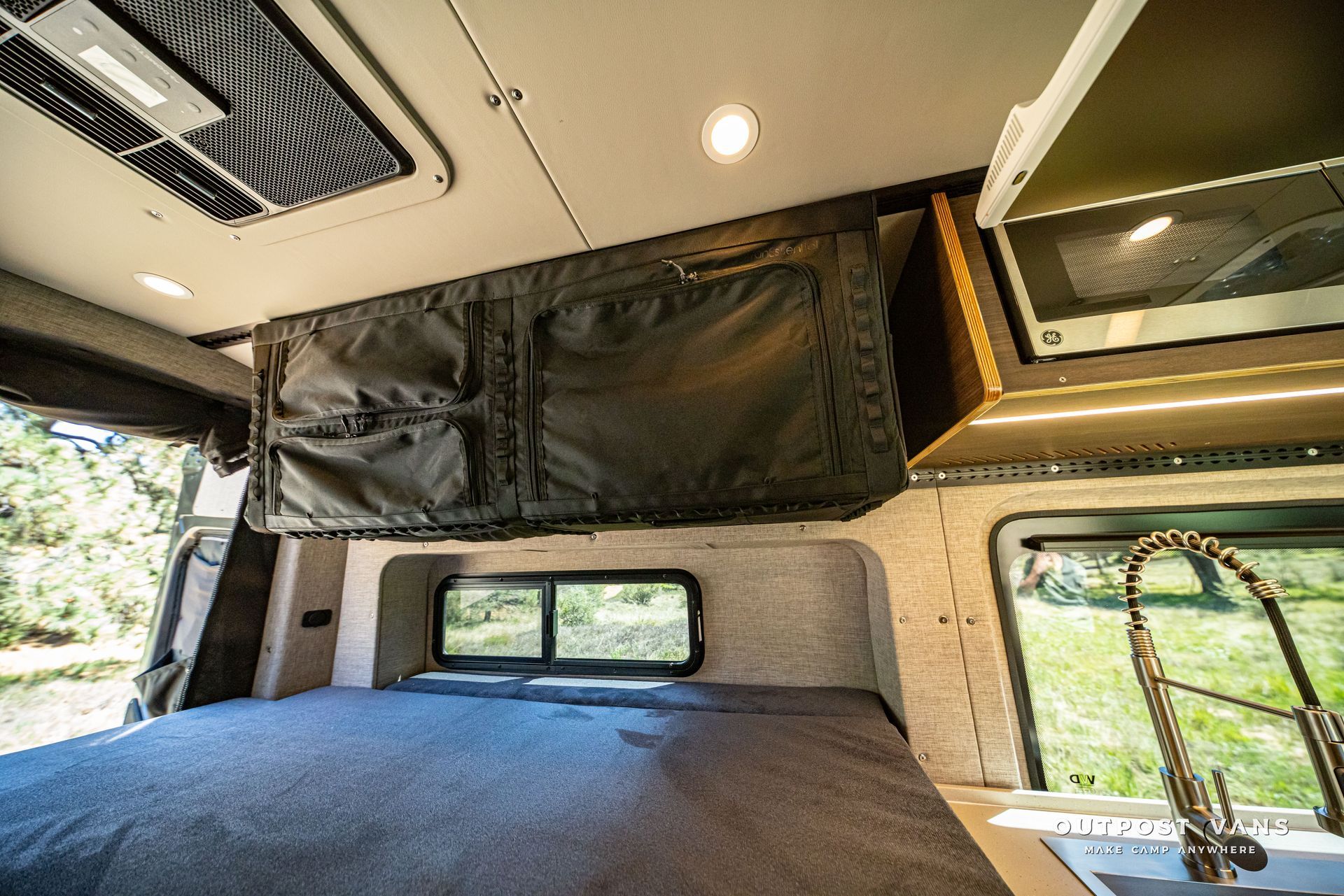 Inside a camper van, a black bunk bed, ceiling vent, and window over a gray mattress.