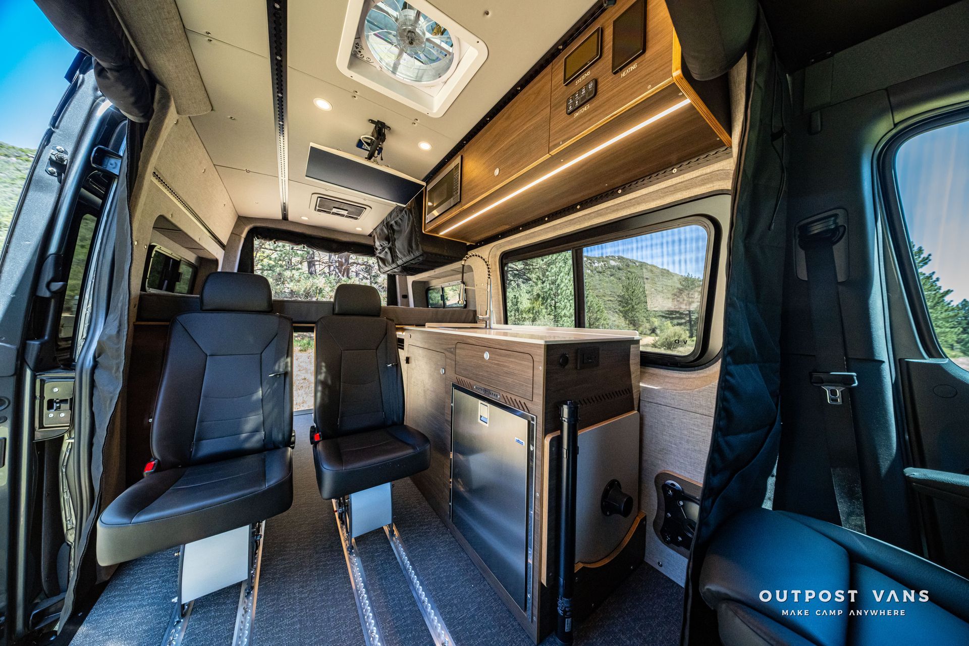 Custom camper van interior with black seats, wood cabinetry, skylight, and window views of trees.
