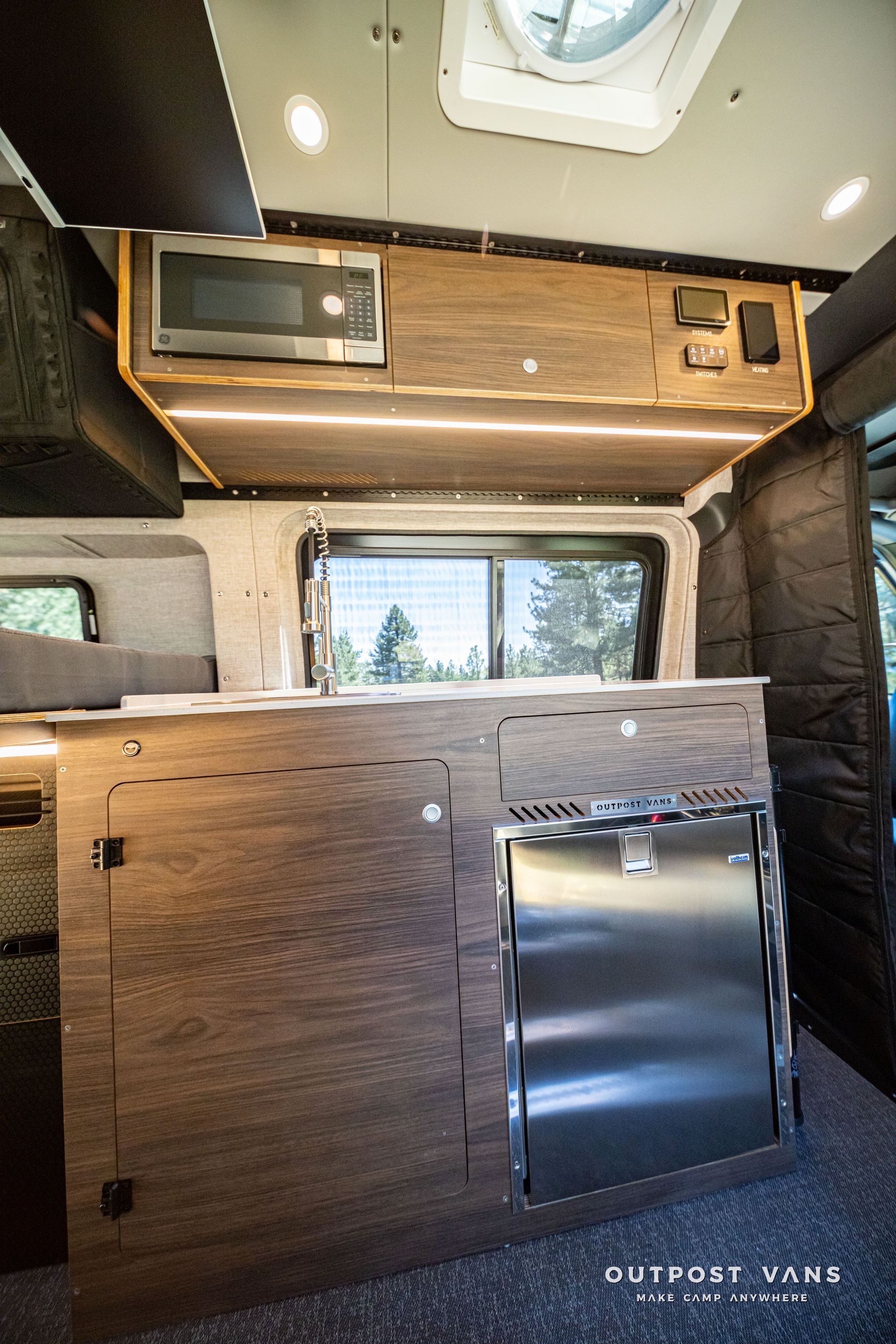 Compact camper van interior with wood cabinets, a window, and a sink under a skylight.