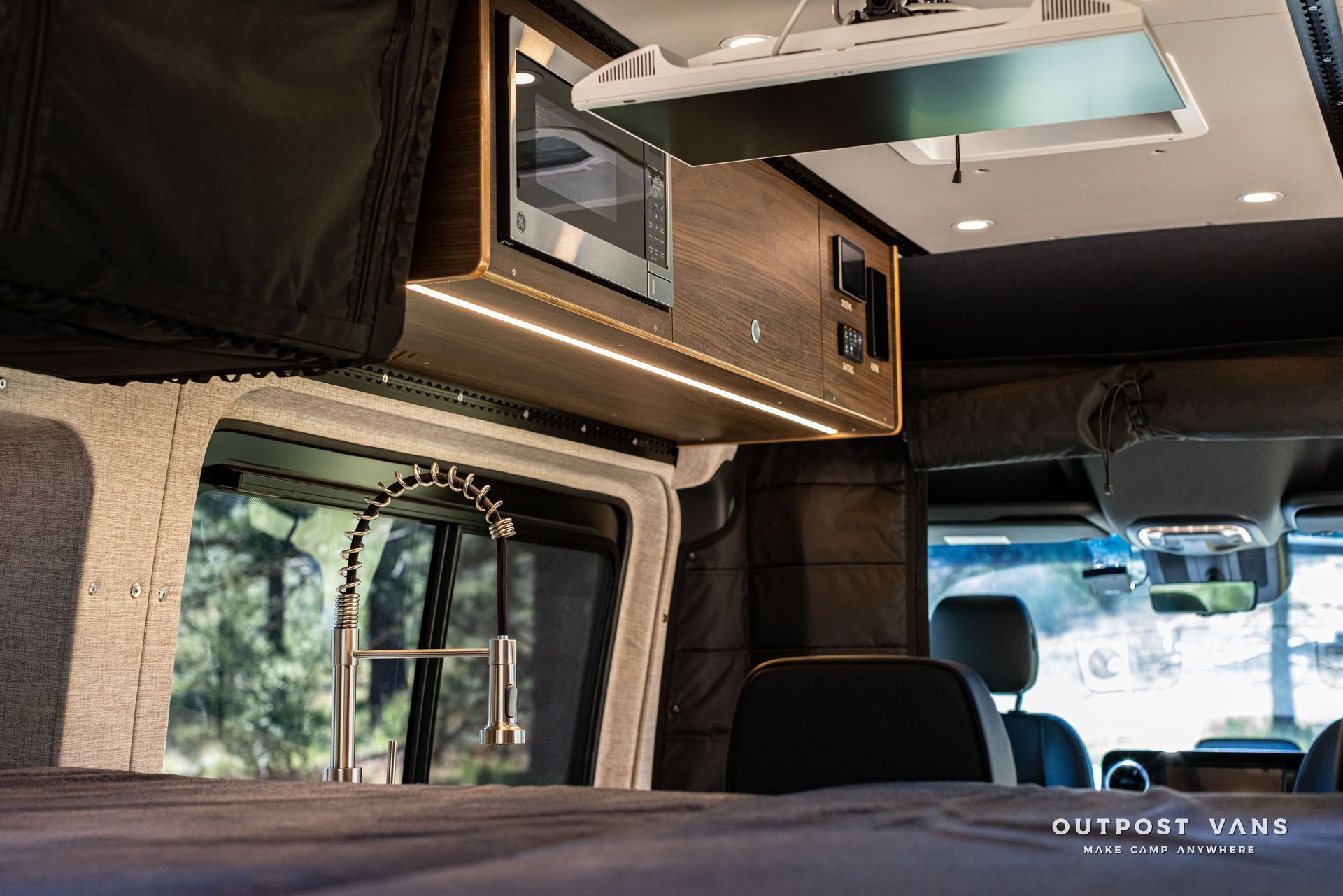 RV interior with bed, overhead storage, and window view of trees and a driveway