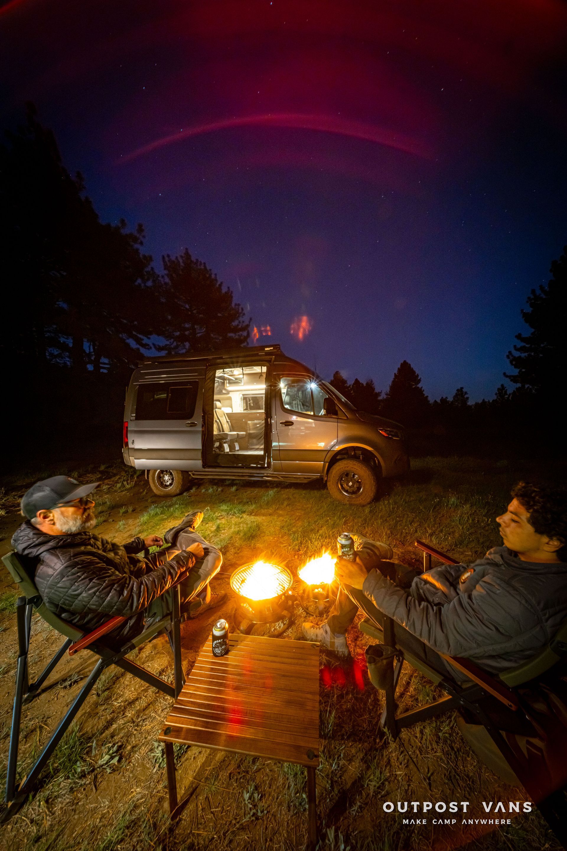 Two campers sit by a glowing campfire beside a van under a purple night sky