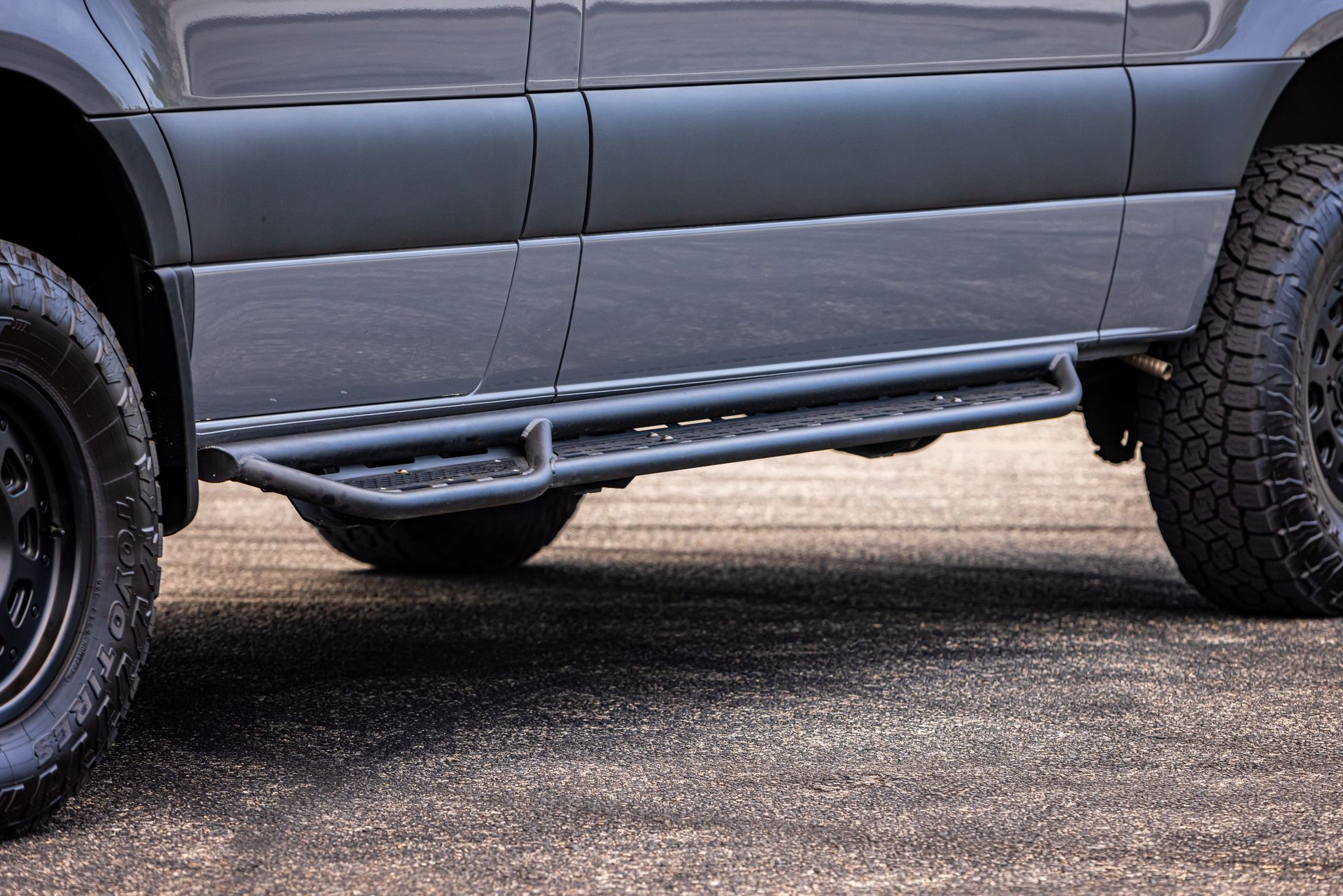 A gray van with black tires and side steps is parked on the side of the road.